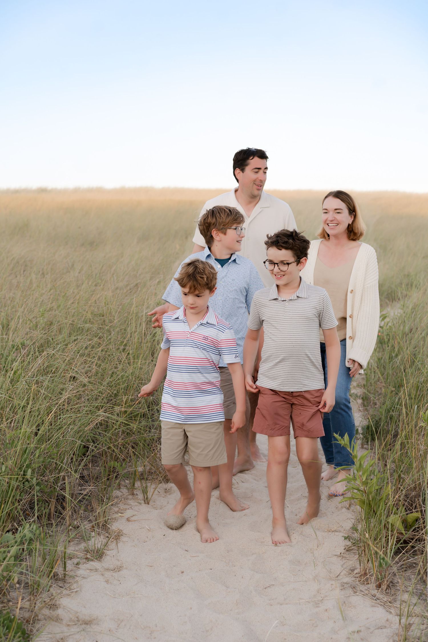 Family walking together on sandy beach path through tall grass.