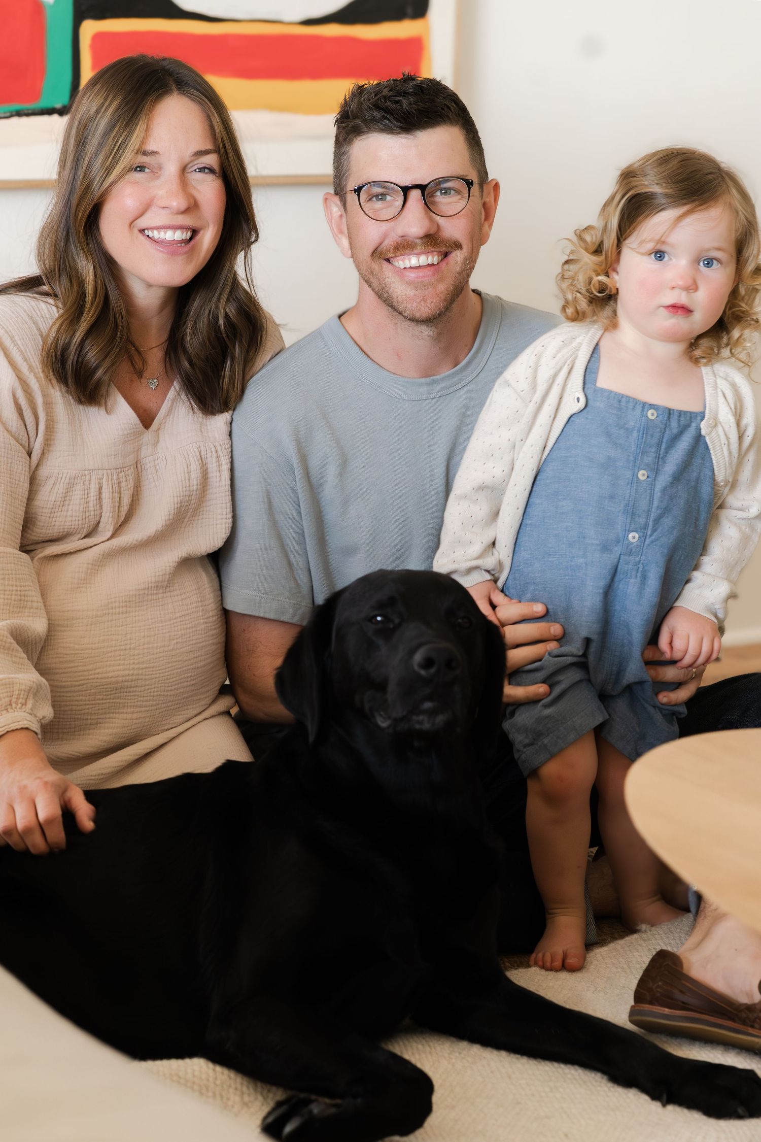 Family sitting together on couch with their black dog beneath colorful abstract artwork.