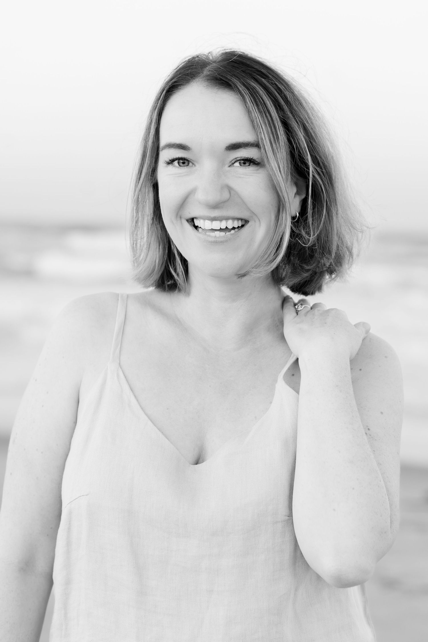 A black and white portrait of a woman smiling at the beach wearing a sleeveless top.