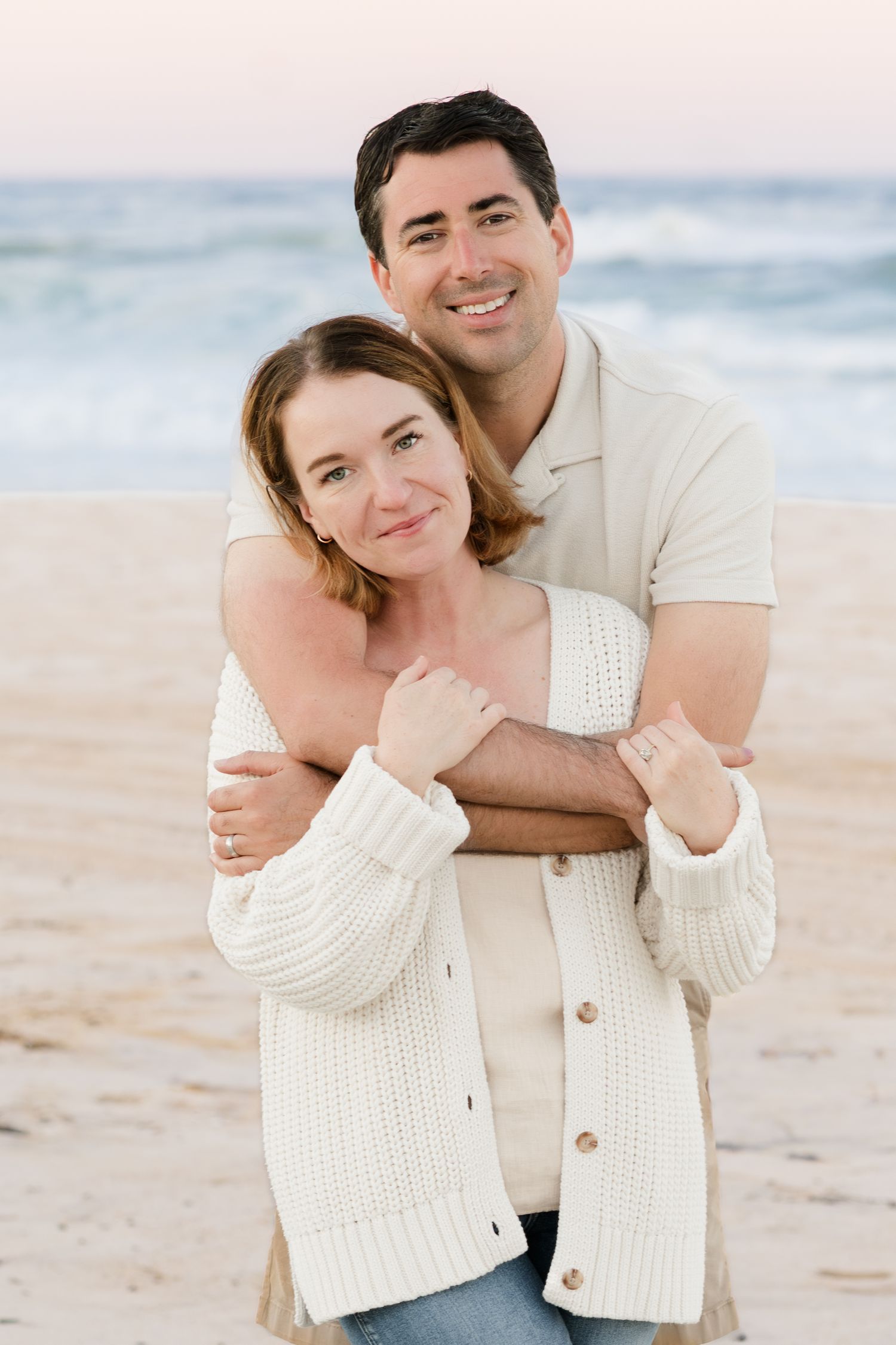 Couple embracing on beach wearing white sweaters at sunset.
