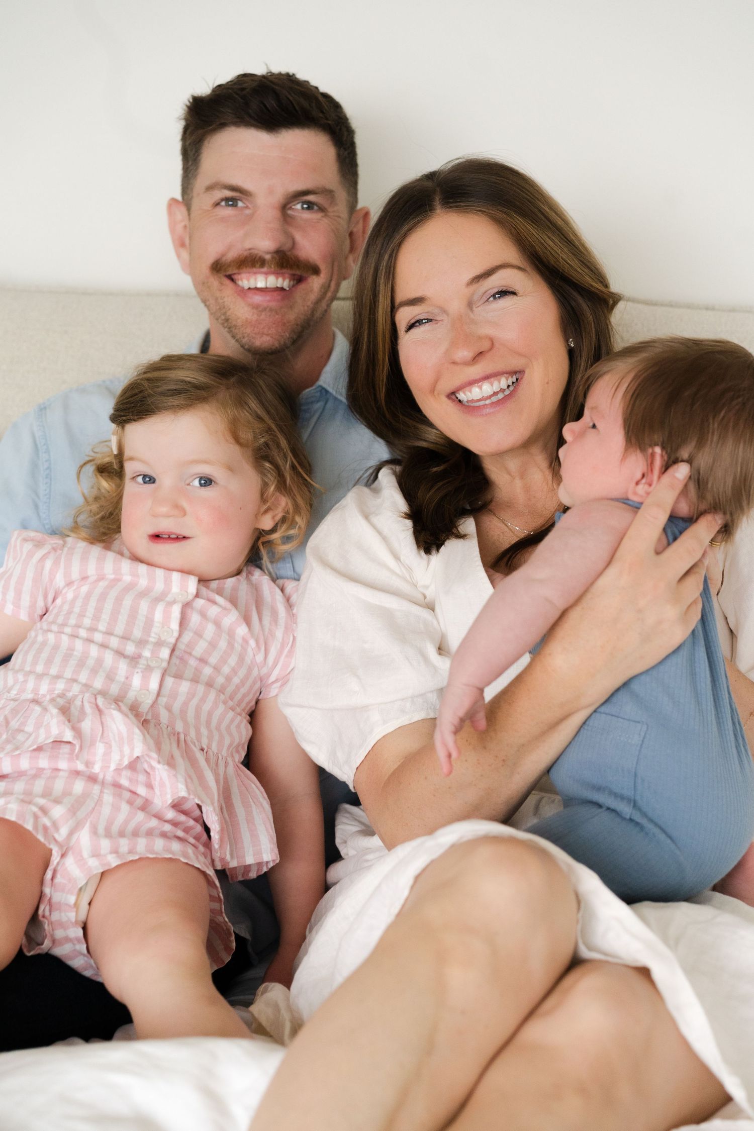 Family cuddles together on white bedding during casual lifestyle photoshoot.