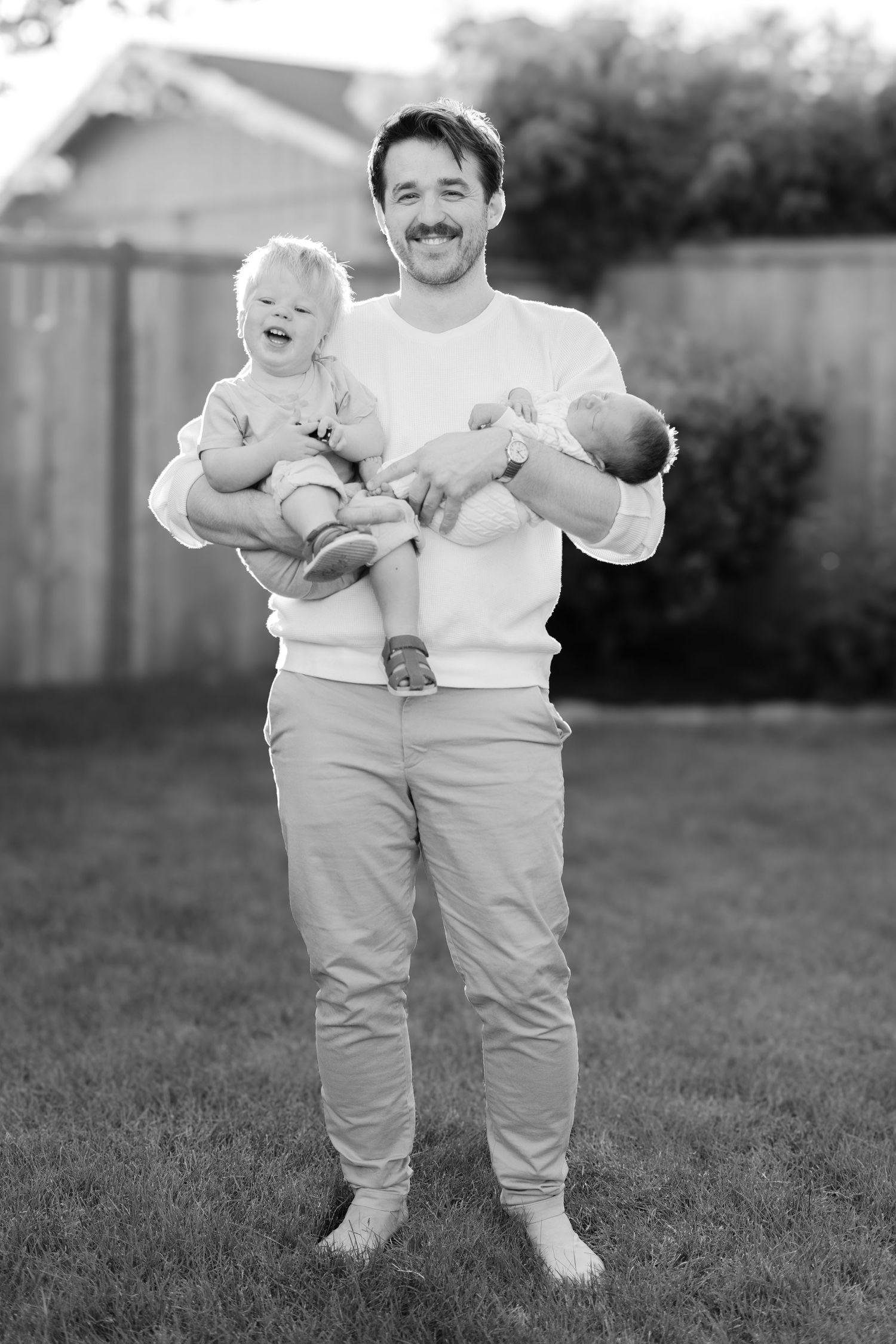 Black and white outdoor photo of person holding both a toddler and newborn.