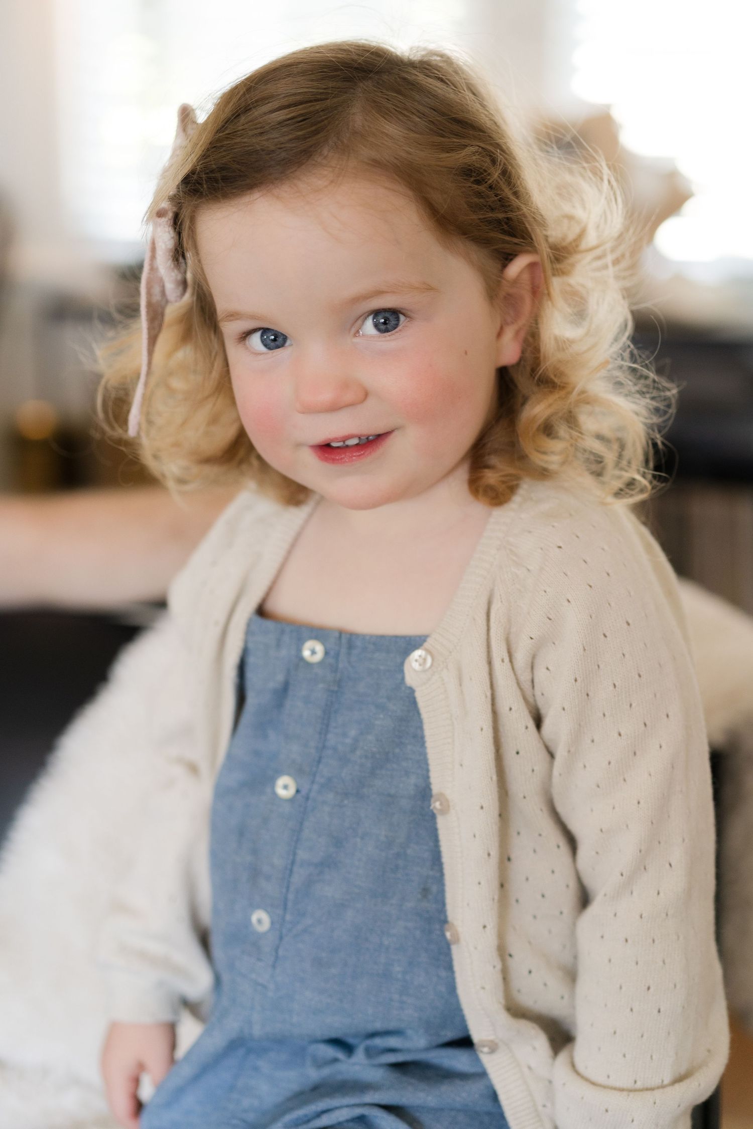 Toddler wearing blue overalls and beige cardigan smiles while sitting.