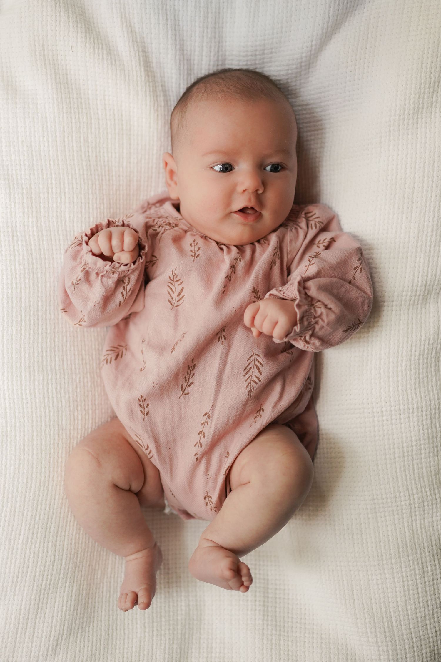 Baby wearing pink floral romper with puff sleeves lying on white textured blanket.