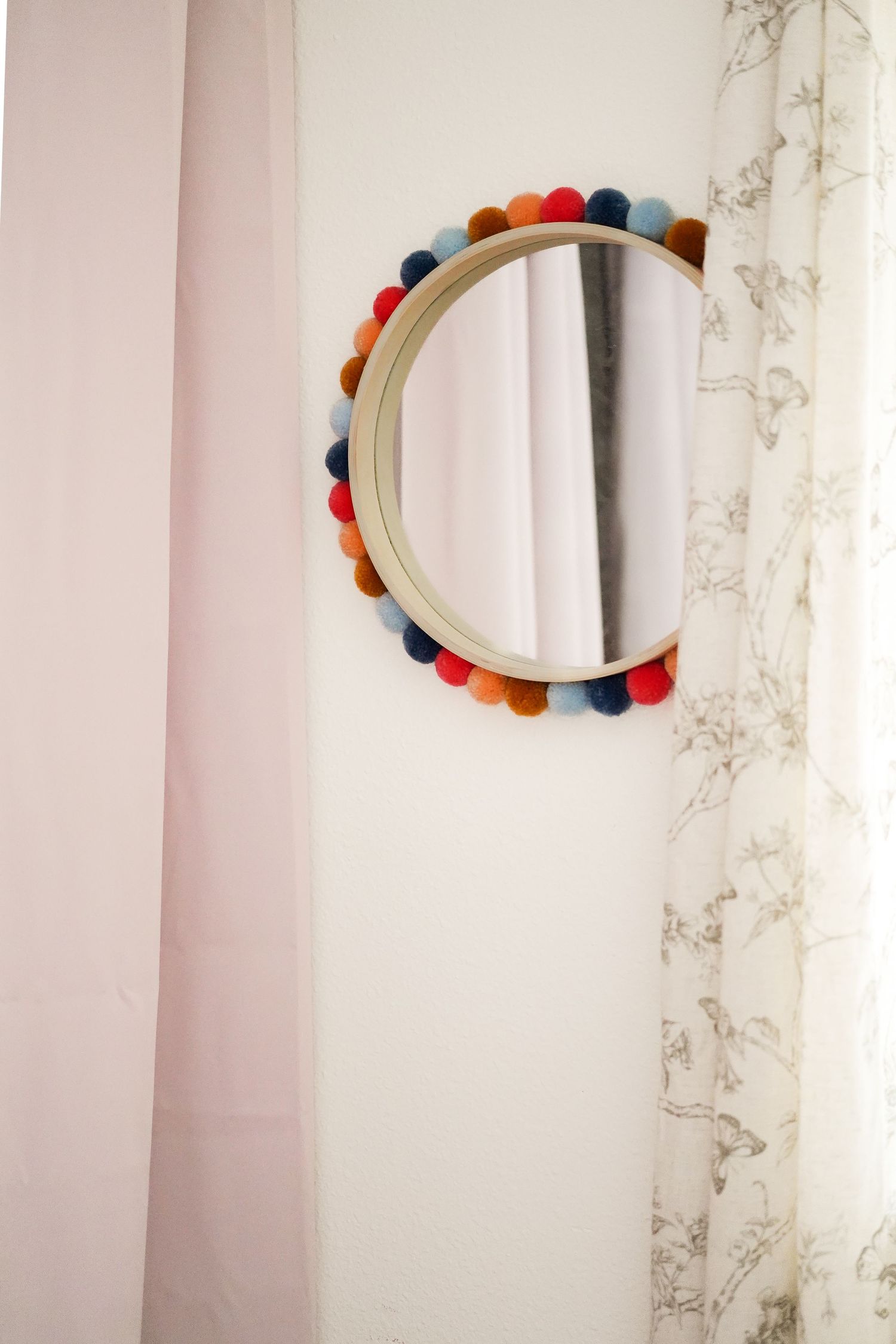 Round mirror decorated with colorful pom poms hanging on pink wall.