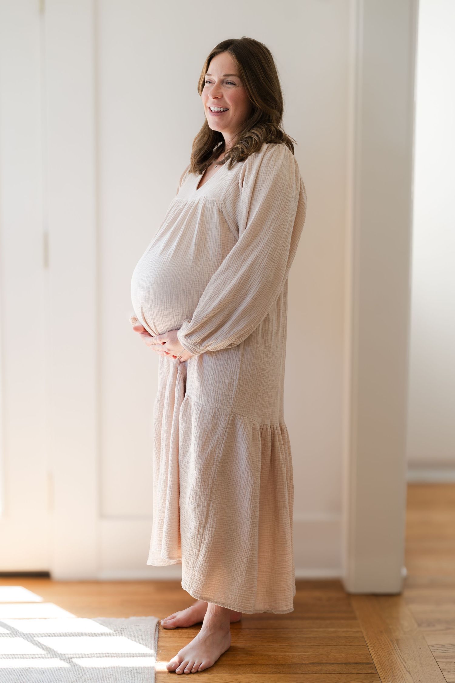 A side view of an expectant parent in a light beige long-sleeved dress standing against a white wall.