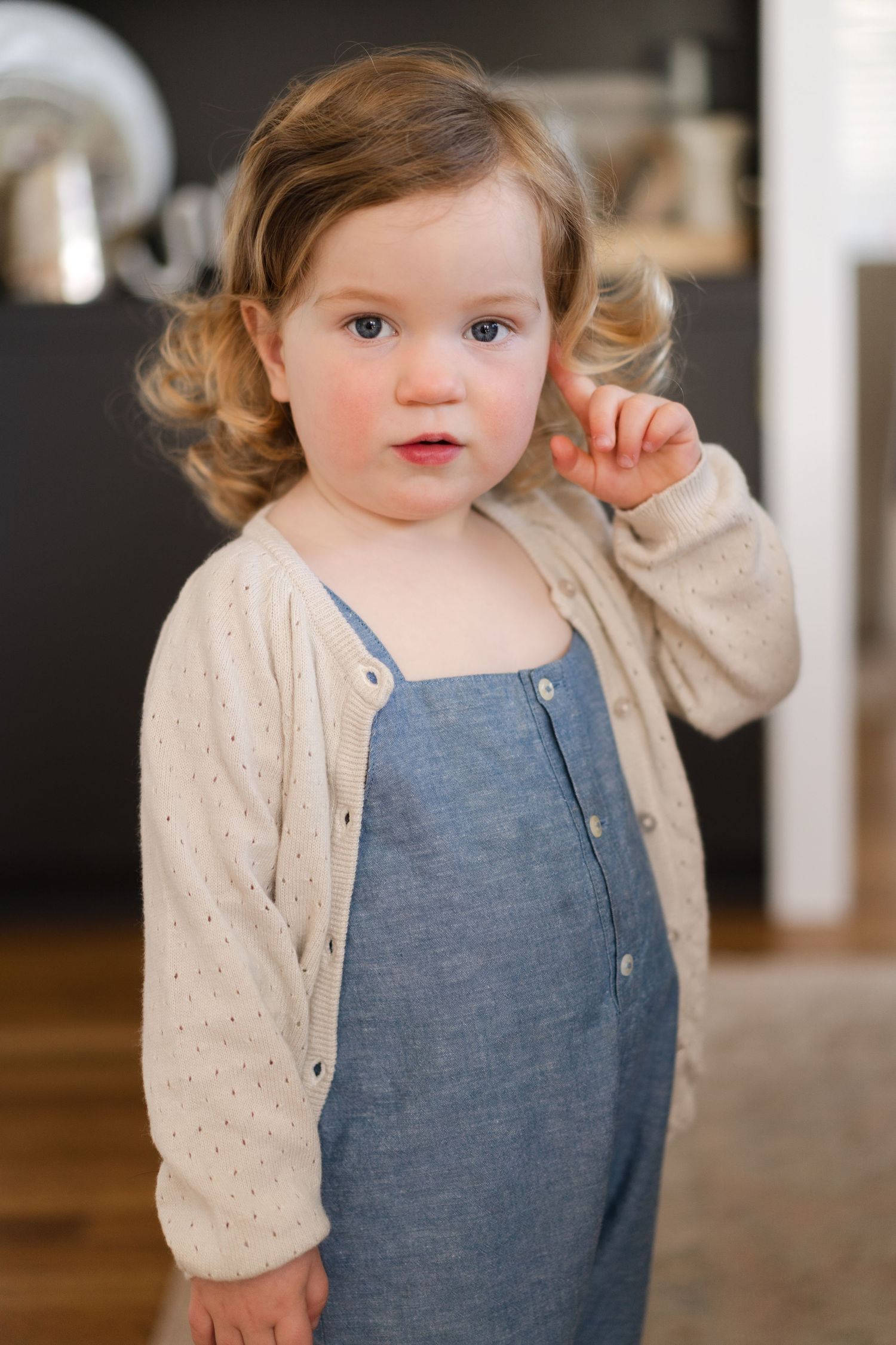 Toddler smiles sweetly while wearing blue overalls and cream colored sweater.