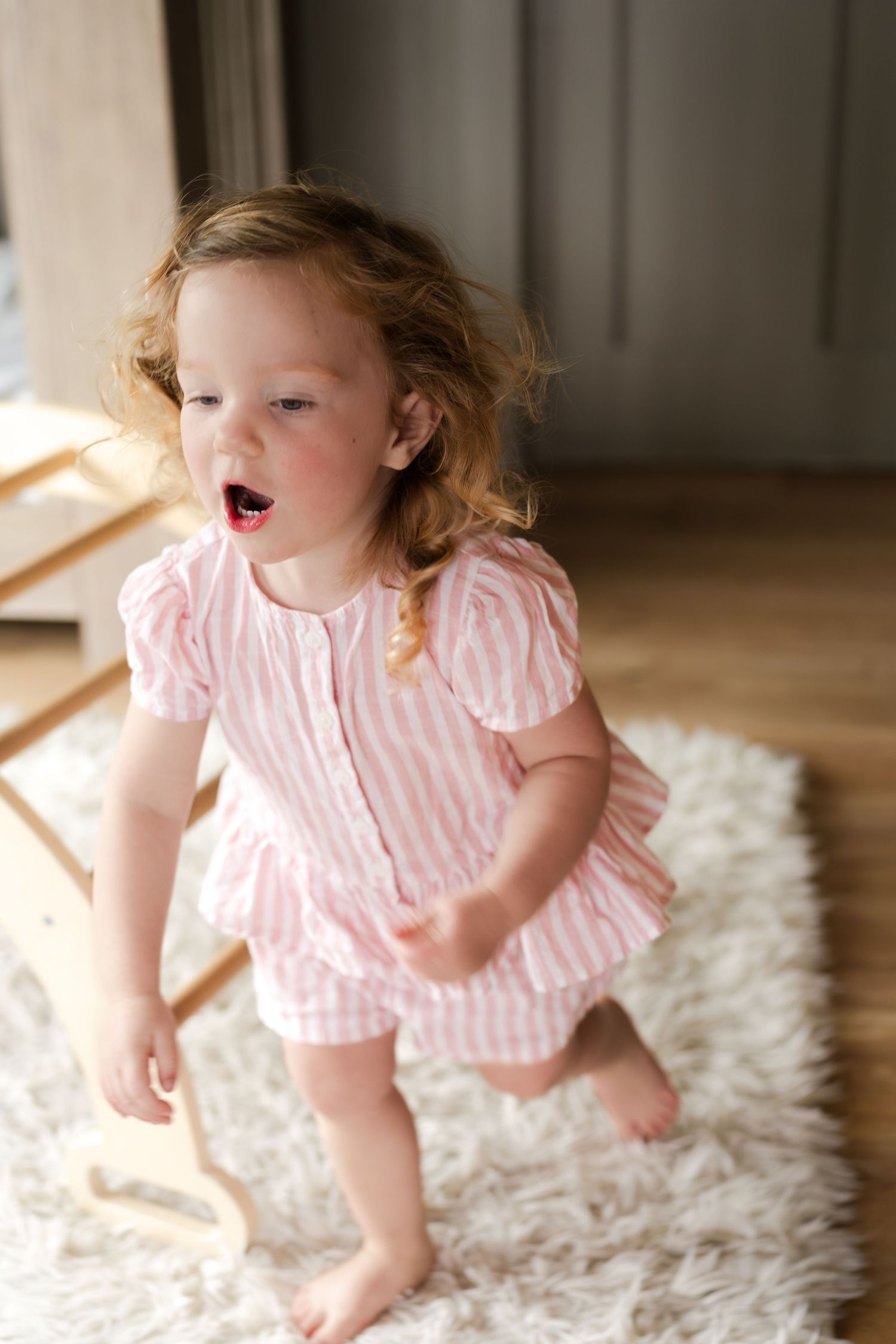 A toddler in a pink striped dress plays on a white fluffy rug in a series of candid shots.