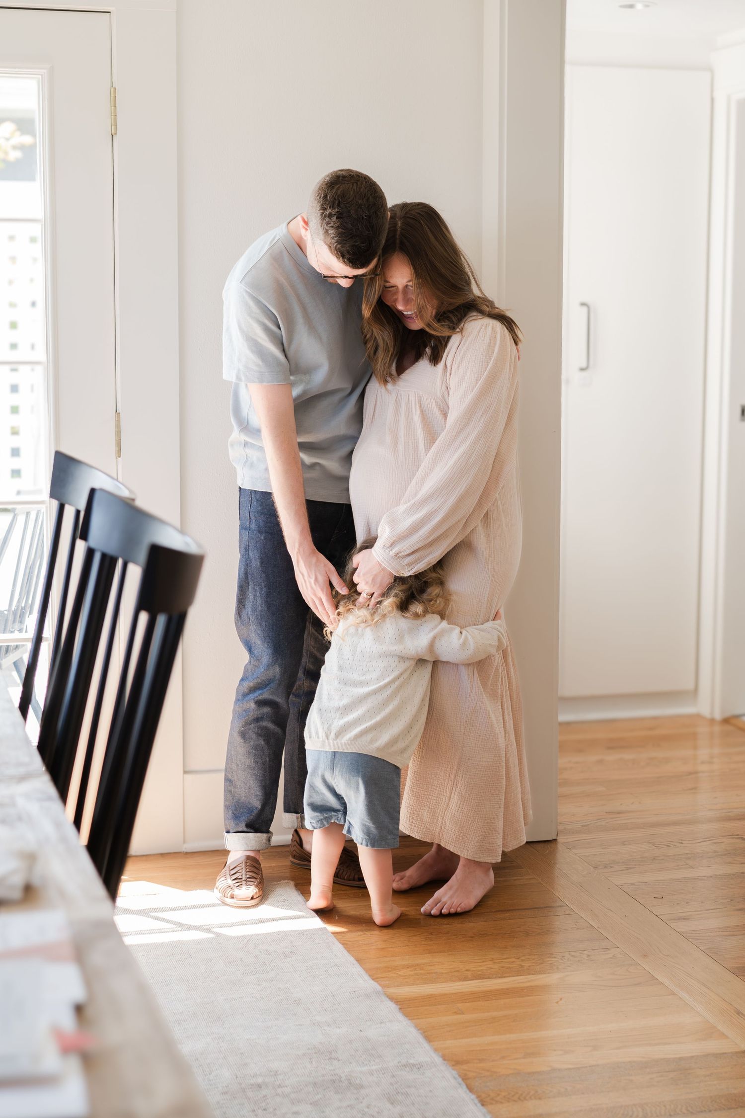 A couple in casual attire stands together intimately on a hardwood floor near a white wall in a bright room.
