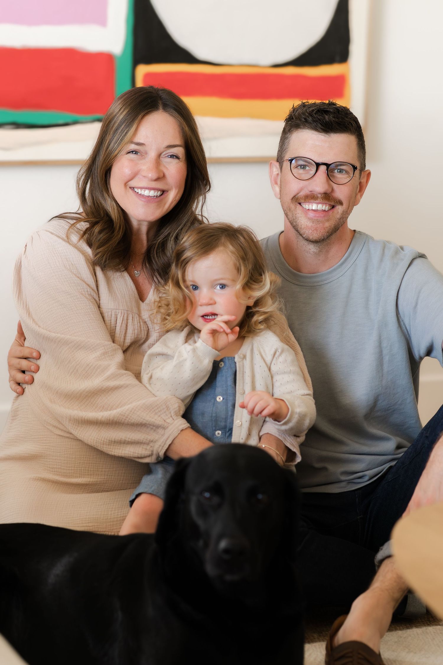 A family sits together on a couch with their black dog in a bright living room with colorful wall art.