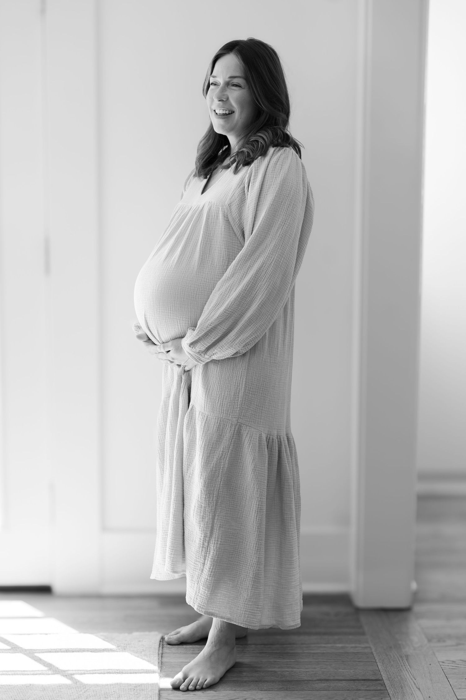A black and white portrait of an expectant parent in a flowing dress with natural window light.