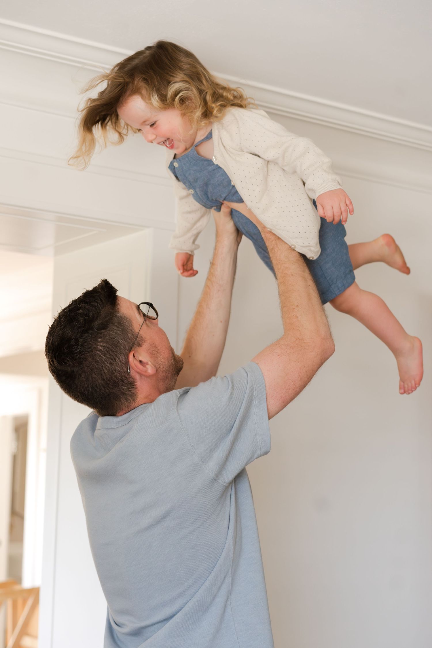 A parent lifts a child wearing denim and cream clothing playfully in the air at home.