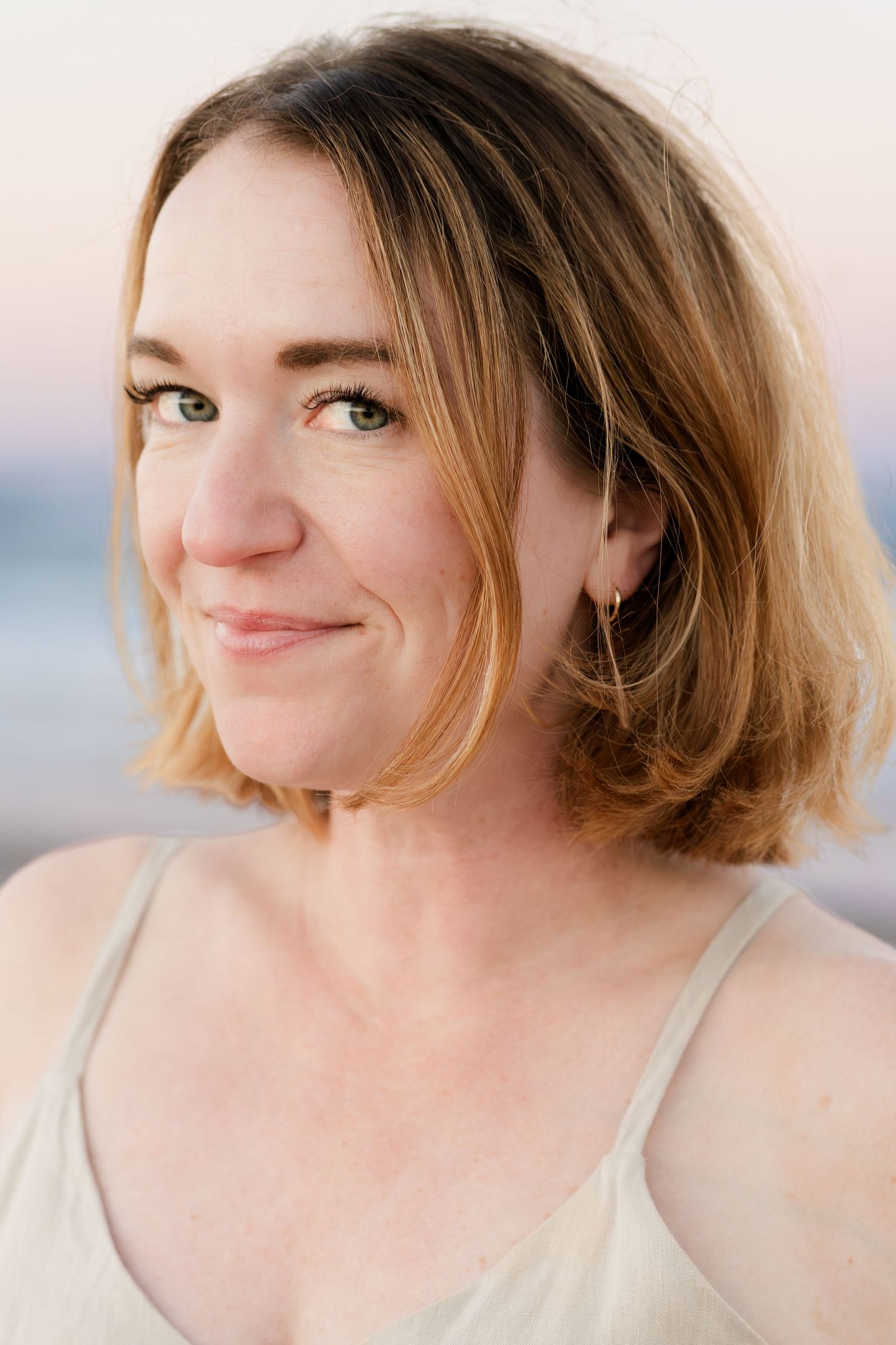 Close-up portrait of someone with shoulder-length blonde hair smiling warmly in soft natural lighting.