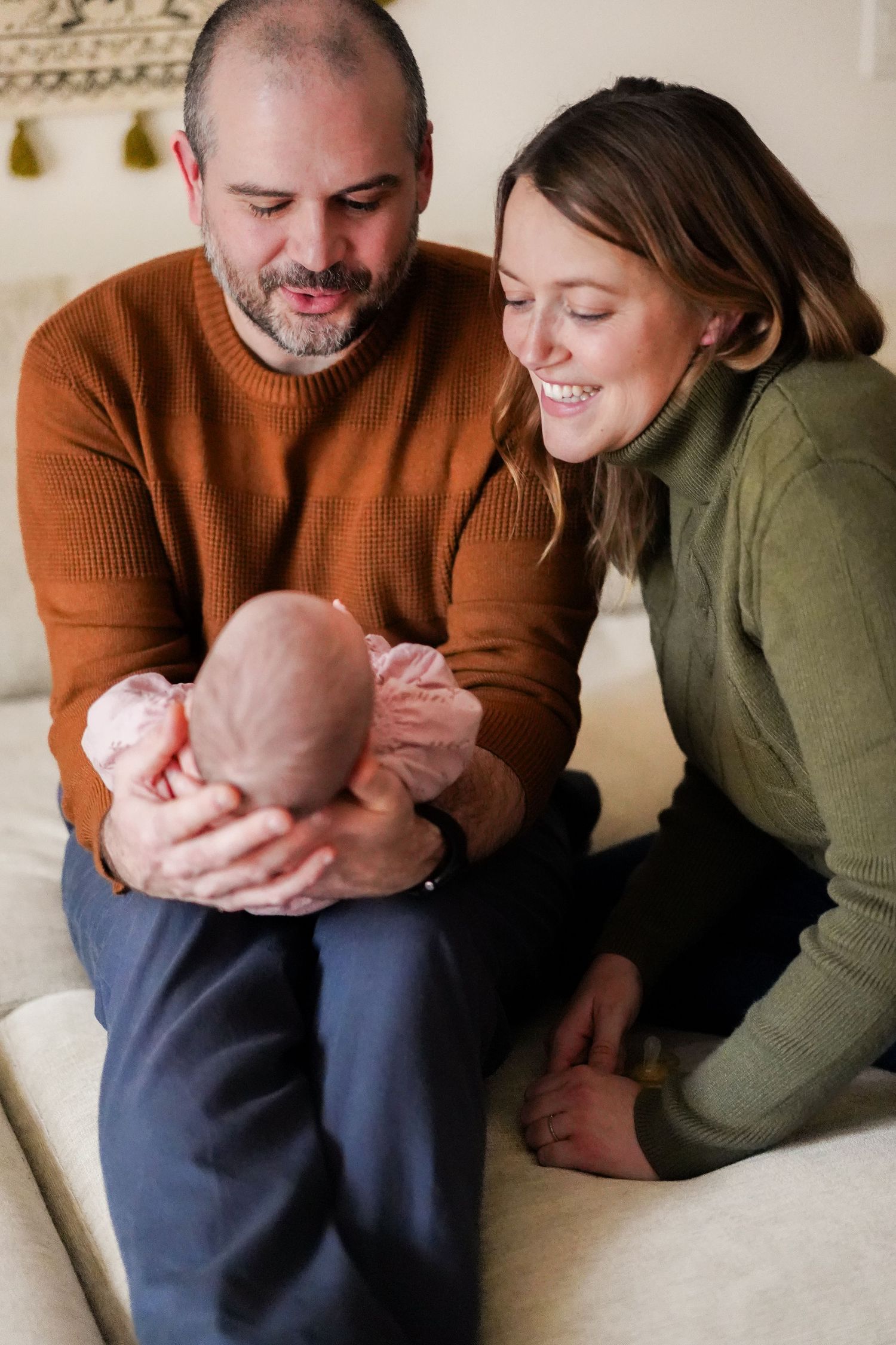 Couple in cozy sweaters sharing tender moment with their baby on light colored couch.