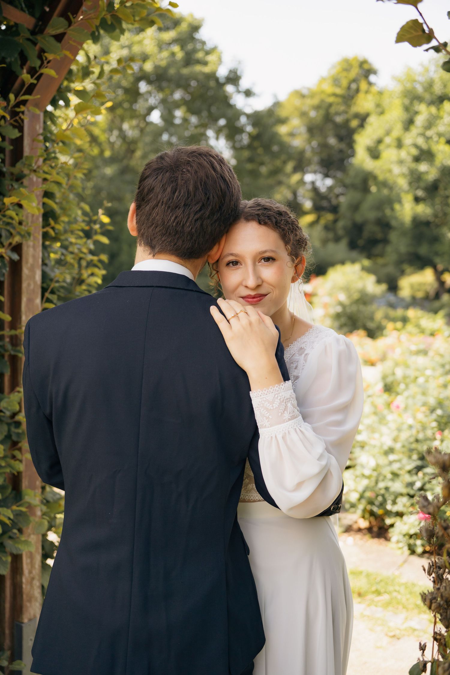 Braut steht hinter dem Bräutigam im Garten, schaut ruhig in die Kamera, intimes Hochzeitsporträt im Grünen