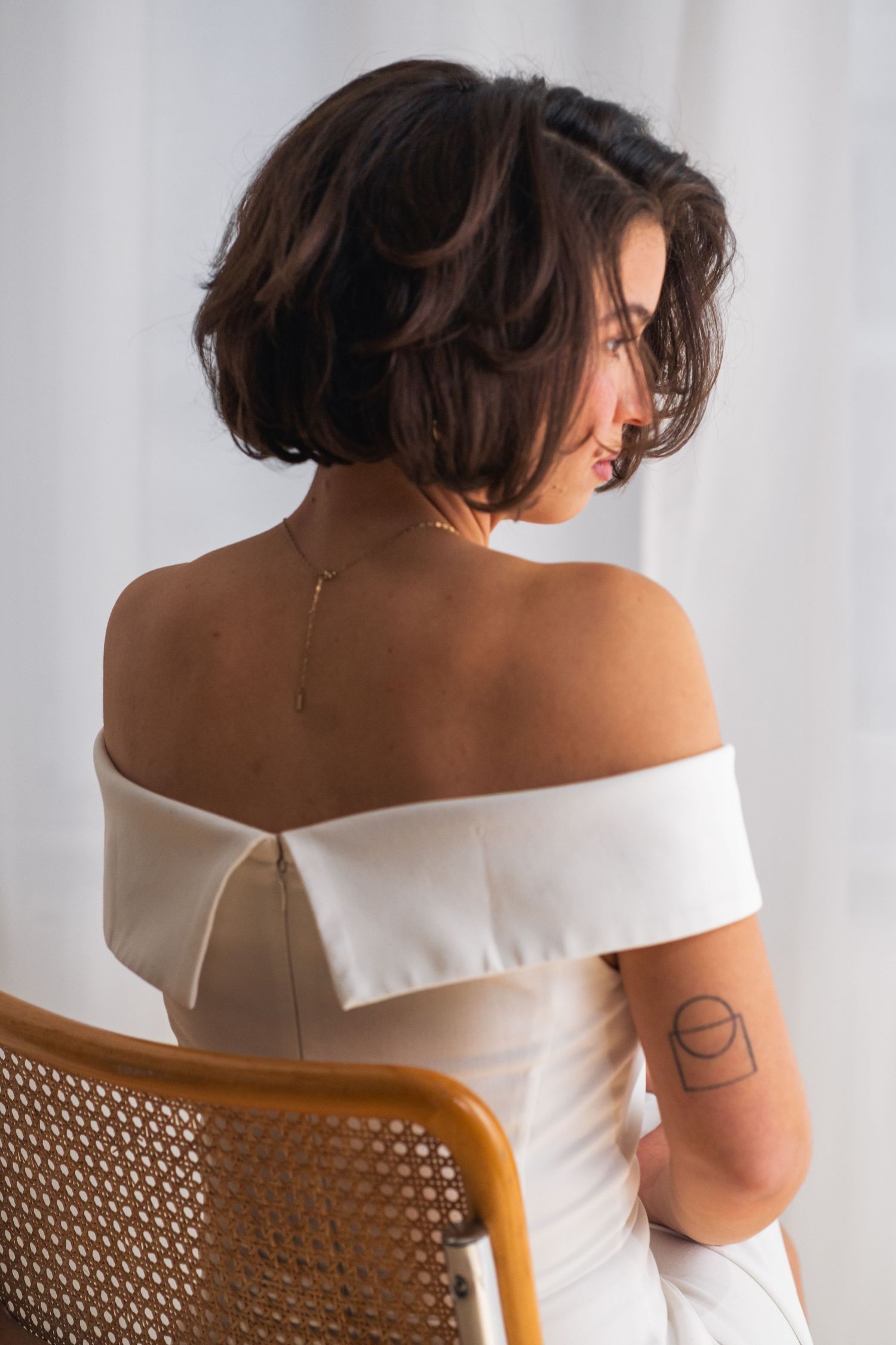 Braut sitzt leicht gedreht auf einem Stuhl, Rückenansicht im schlichten Brautkleid, ästhetisches Brautporträt