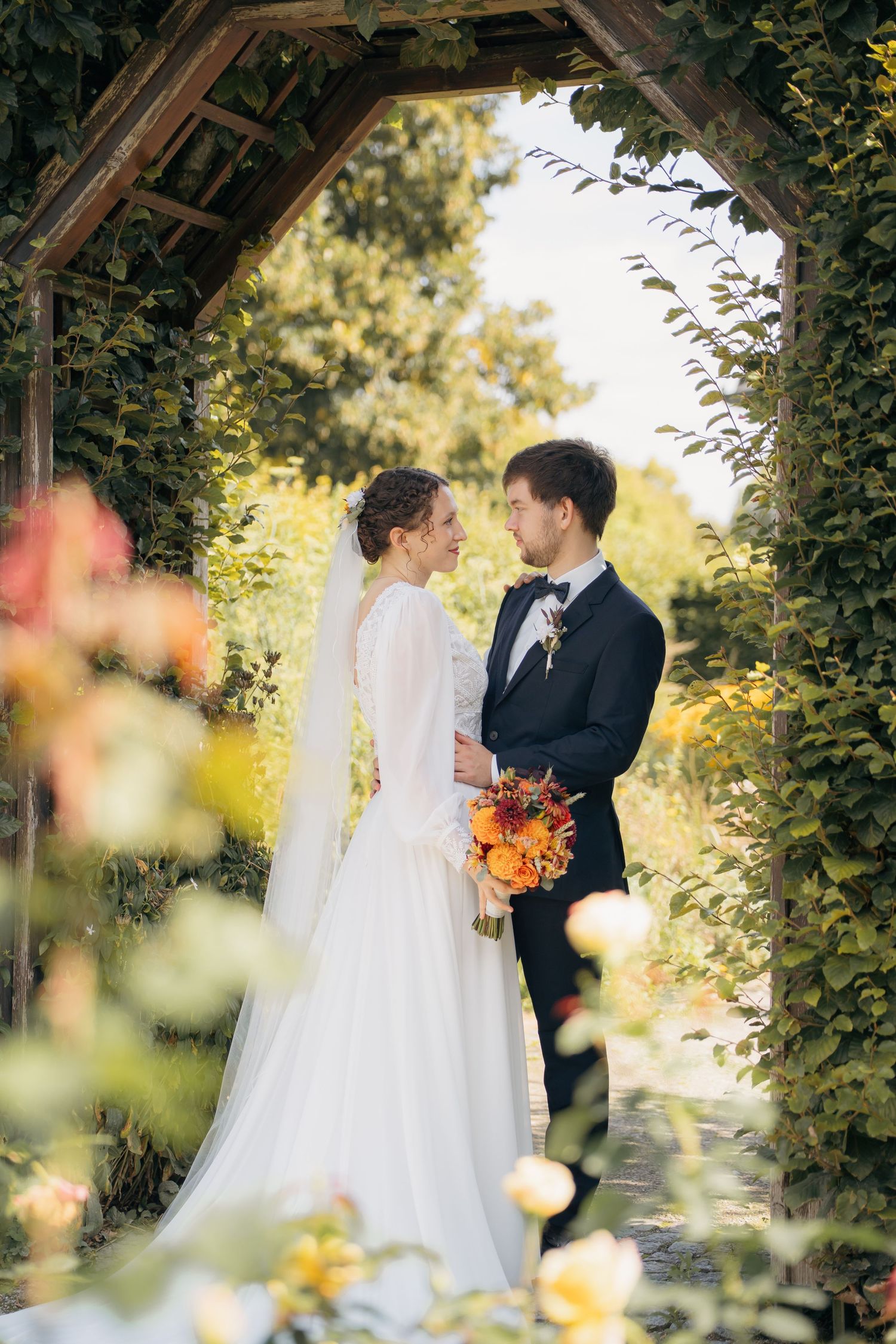 Brautpaar steht sich unter einem bewachsenen Torbogen gegenüber, romantisches Hochzeitsporträt im Garten