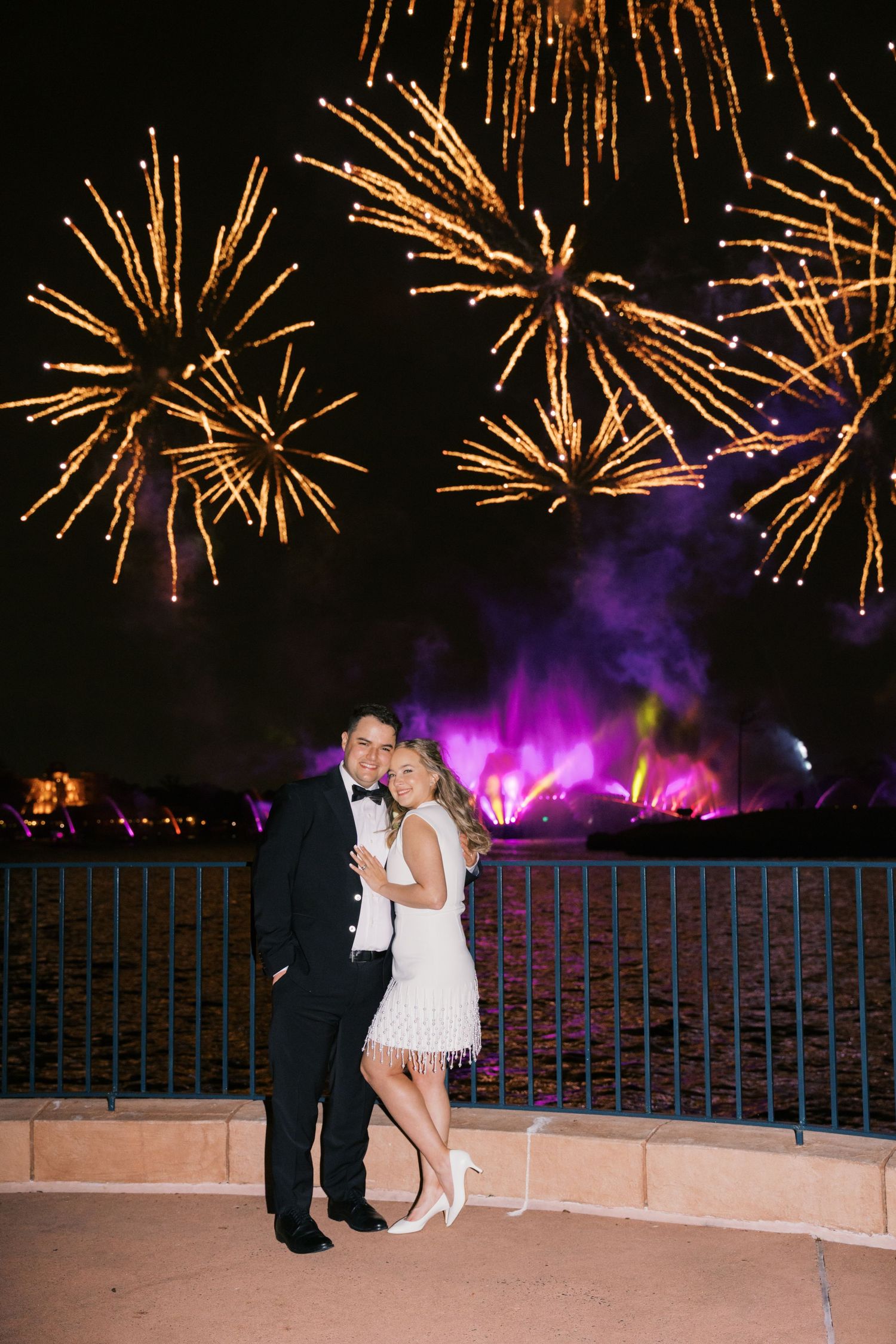 Couple in formal wear poses in front of stunning fireworks display over water at night celebration.