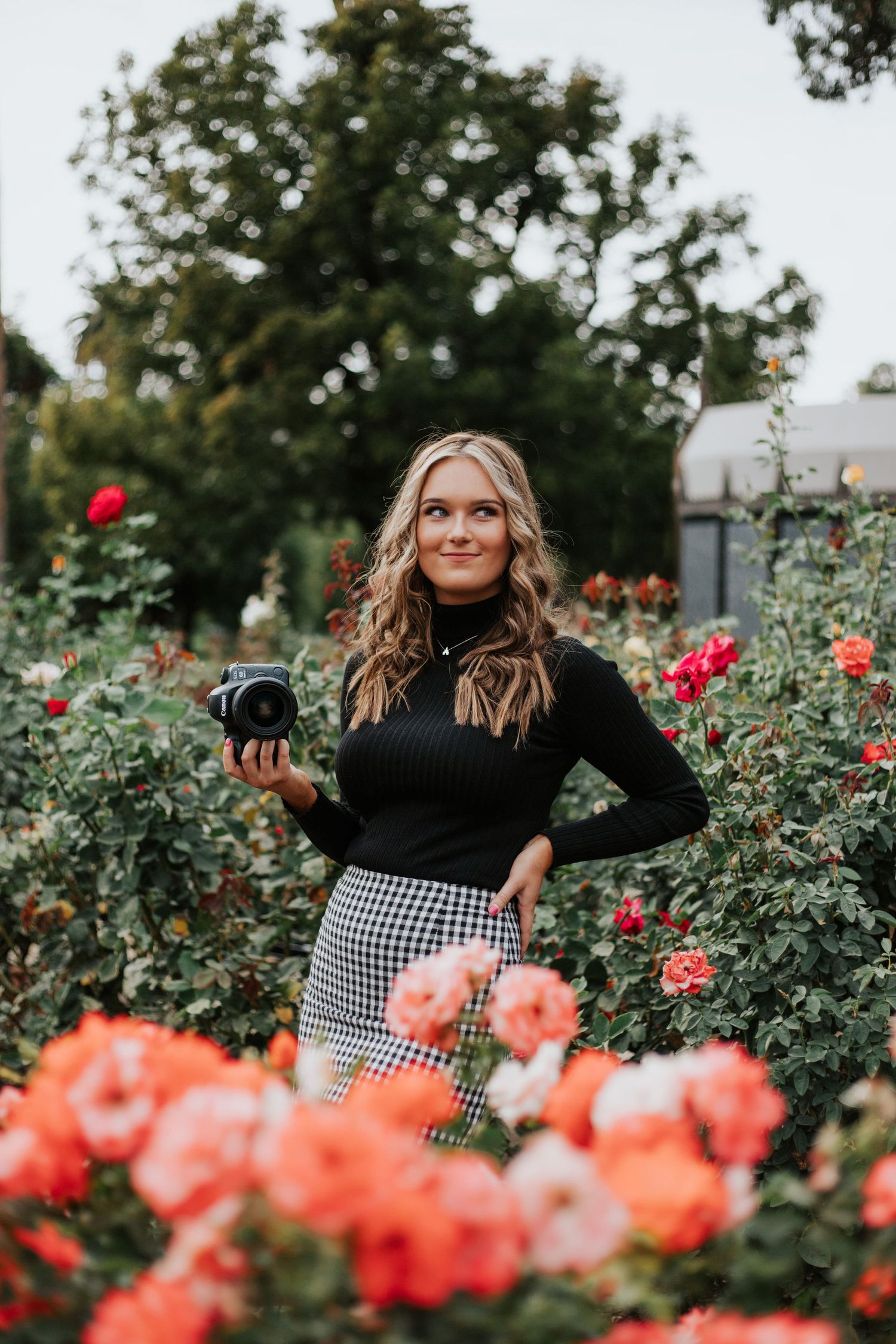 Photographer in black turtleneck and checkered skirt stands among blooming coral roses in garden.