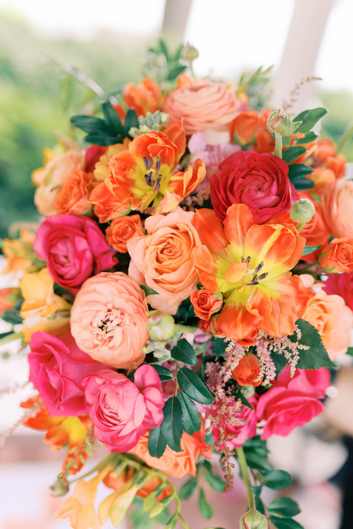 Vibrant wedding bouquet featuring coral, peach and hot pink roses with orange lilies and greenery accents in soft focus.