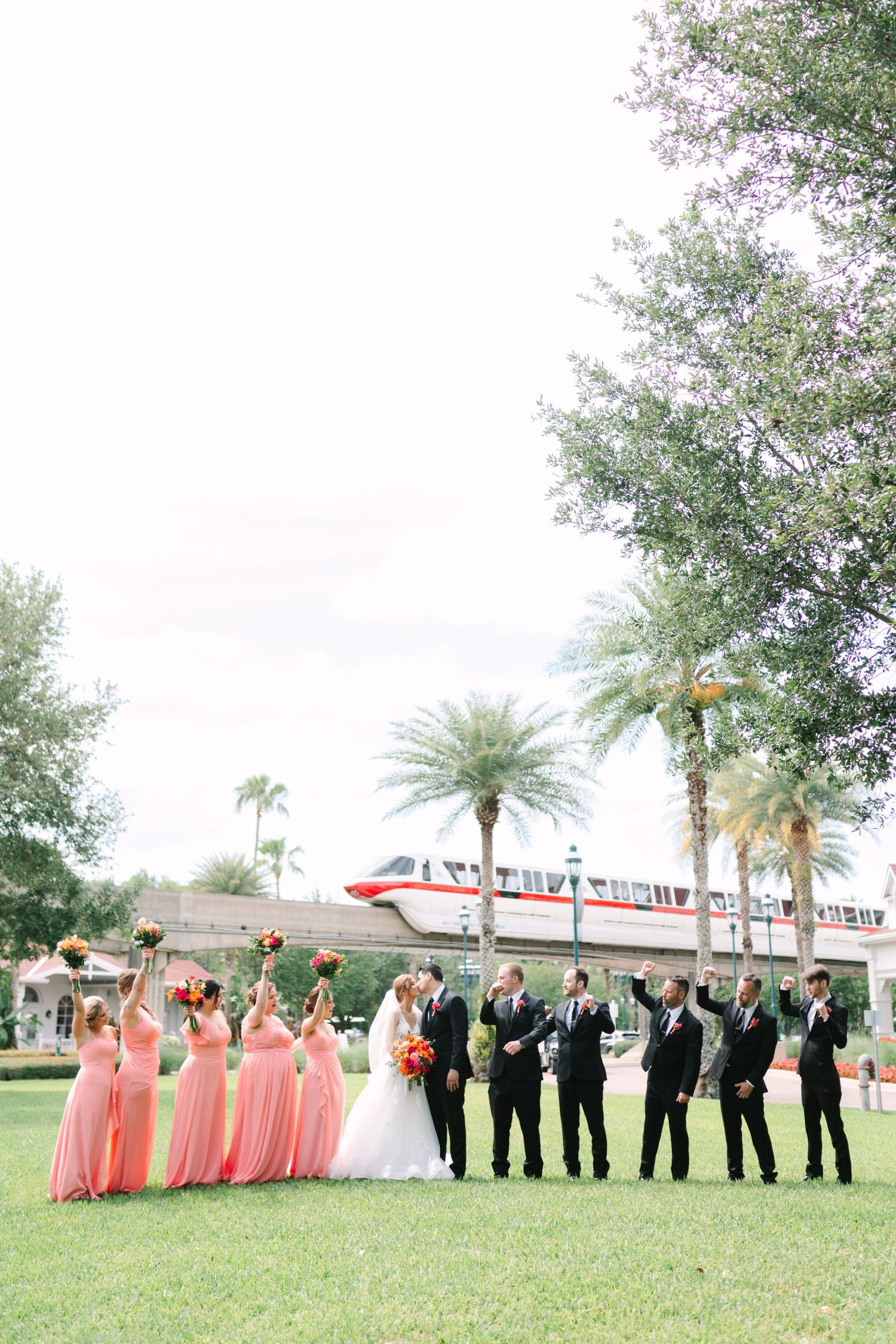 Wedding party poses on green lawn with Disney World monorail and palm trees in background at Disney resort.
