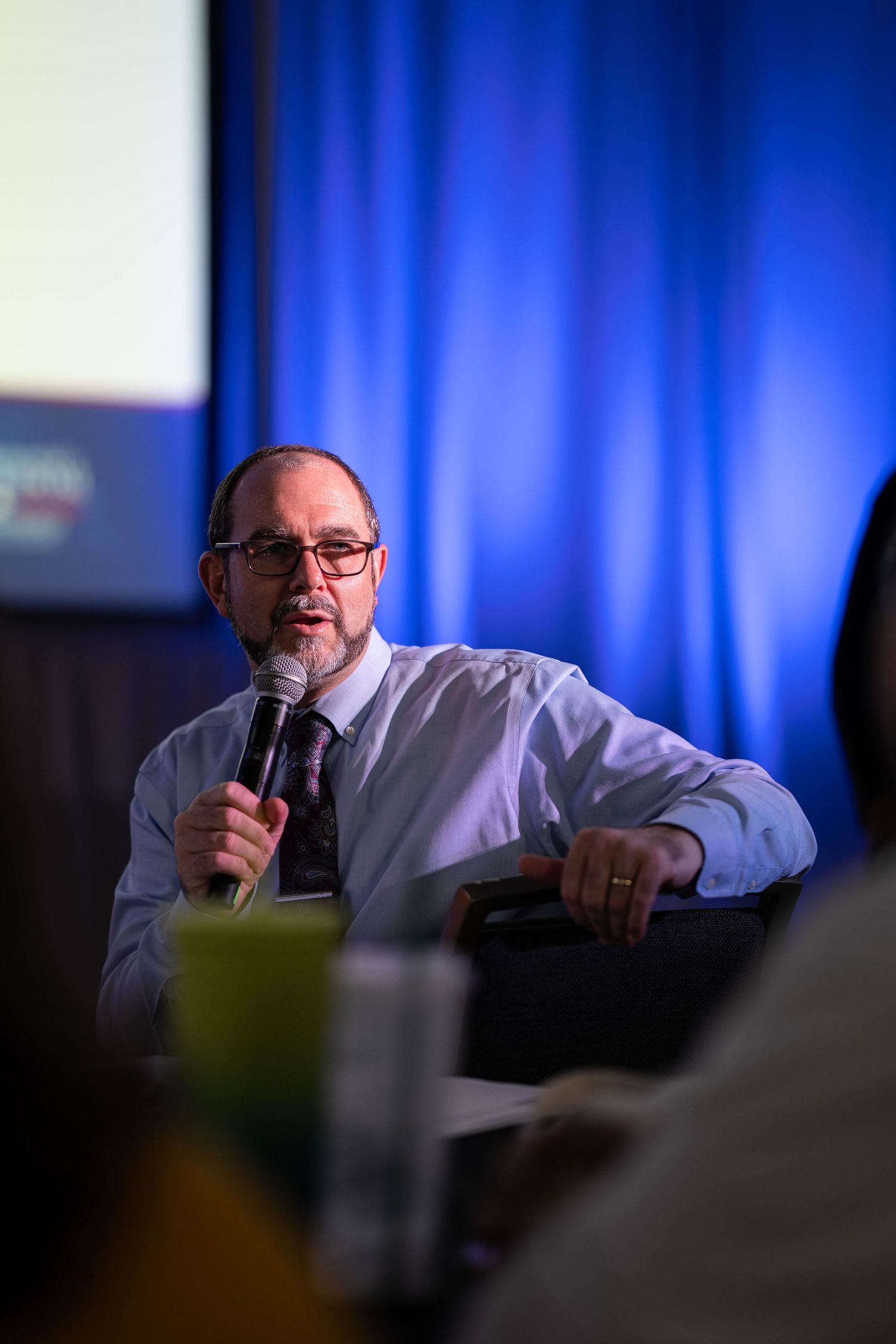 Man with glasses speaking into microphone at conference panel, blue curtain backdrop, professional business setting.