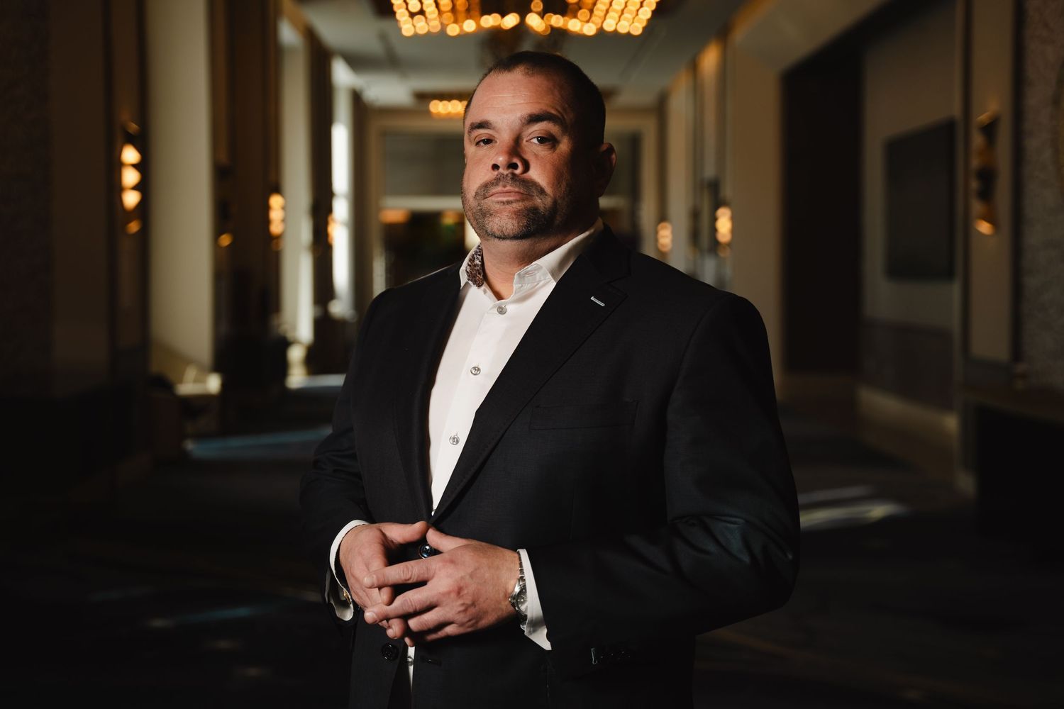Professional man in white shirt and blazer posing confidently in an elegant dimly lit hallway.