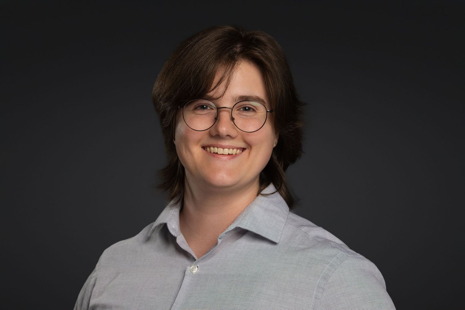 Smiling person with brown hair and glasses wearing a light blue shirt against a dark gray background.