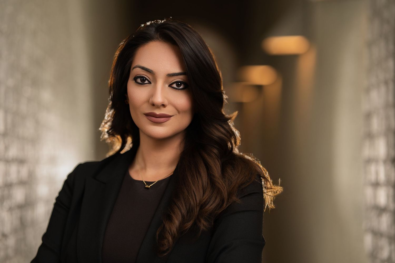 Professional woman in black blazer poses confidently in a softly lit corridor for a corporate portrait.