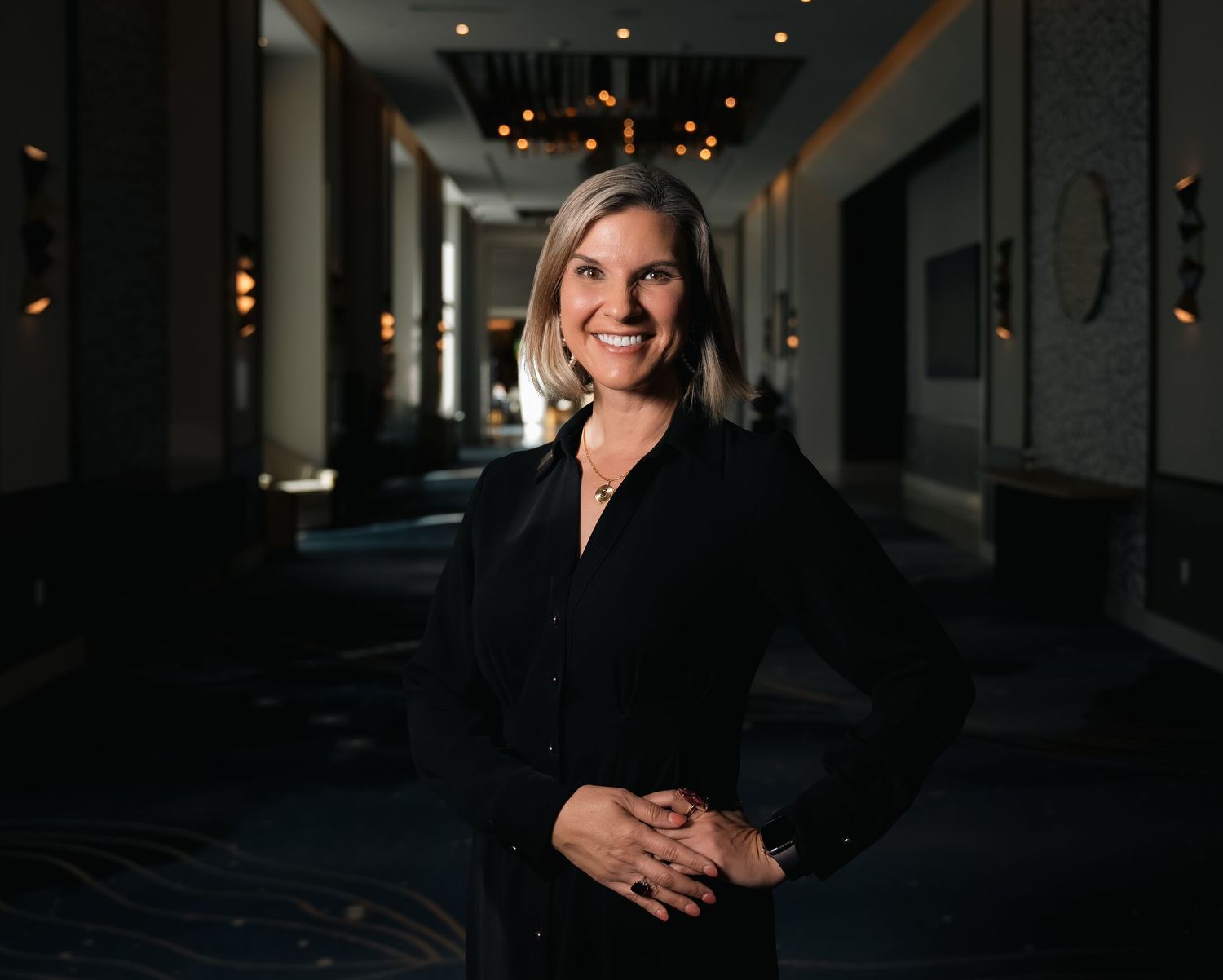 Professional woman in black outfit smiling confidently in an elegant hotel corridor with warm lighting.