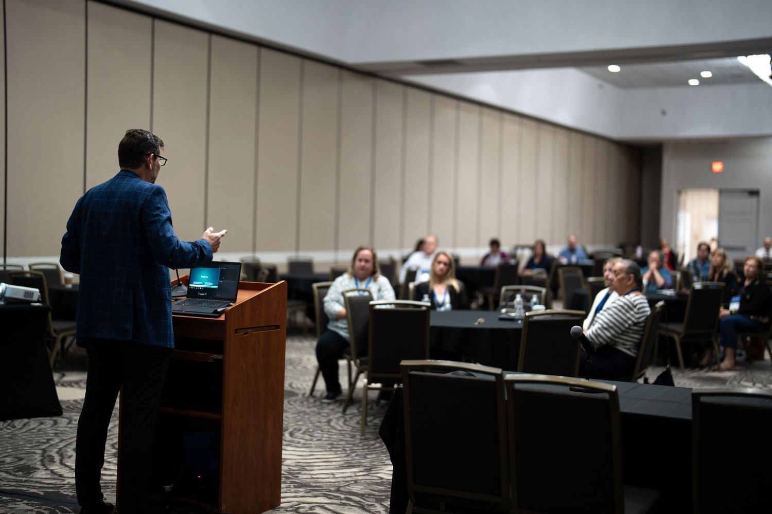 Professional speaker presenting at a corporate conference to an engaged audience seated at round tables in a hotel ballroom.