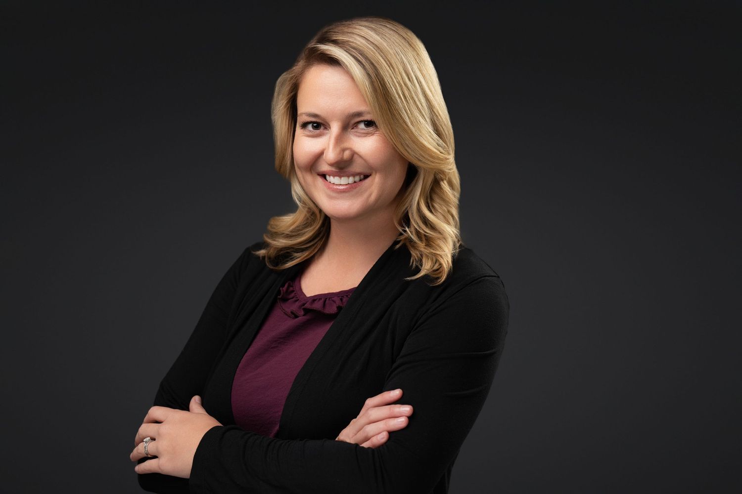 Professional headshot of a smiling blonde woman in a black blazer and maroon top, arms crossed, on dark background.