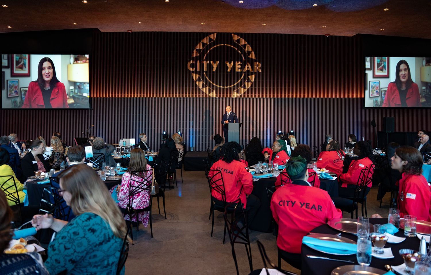 City Year gala event with attendees in red jackets seated at tables, speaker at podium with large screens displaying presenter.