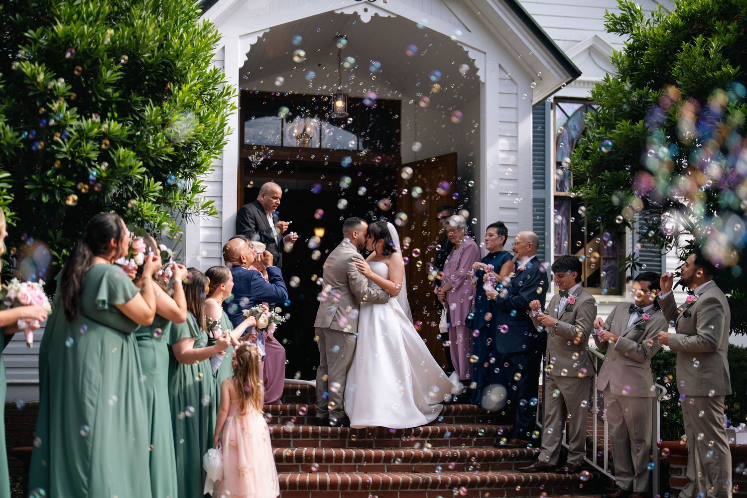 Bride and groom share a kiss on church steps surrounded by bubbles and guests in green bridesmaid dresses.