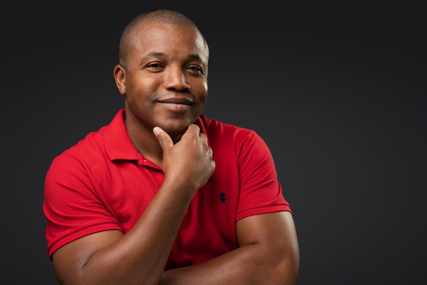 Confident Black man in red polo shirt smiling with hand on chin against dark background.