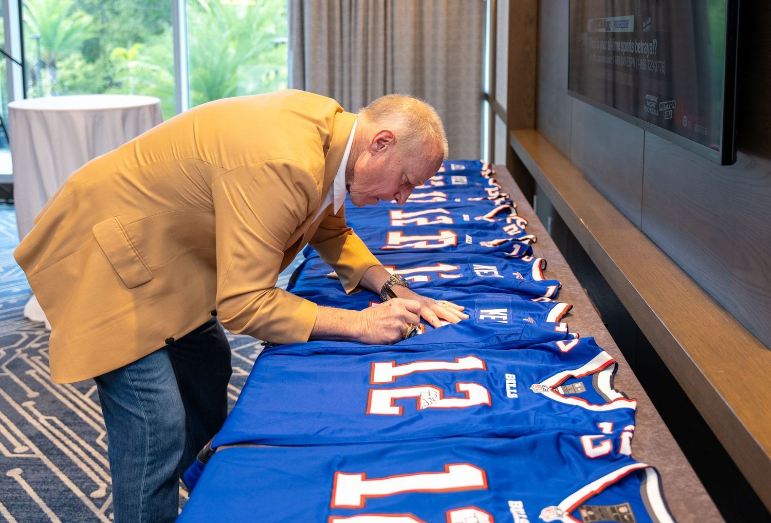 Man in gold blazer signing blue football jerseys displayed on a table at a sports memorabilia signing event.
