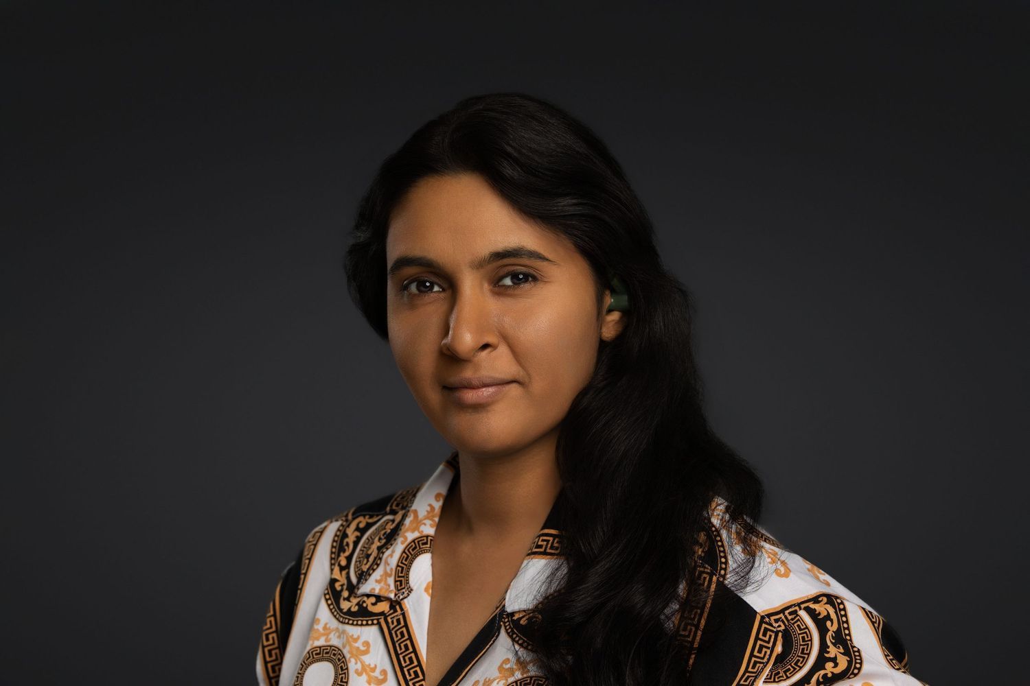 Professional headshot of a young South Asian woman with long dark hair wearing a patterned white blouse against dark background.