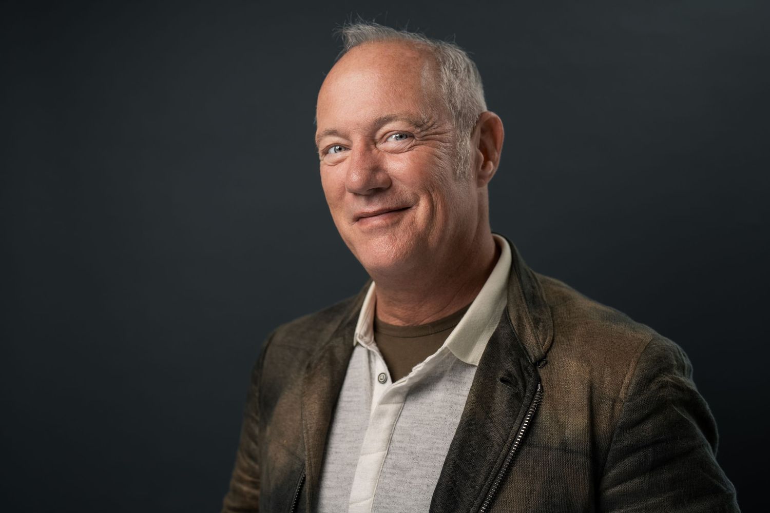 Smiling middle-aged man with gray hair wearing a brown blazer poses for a professional portrait photo.
