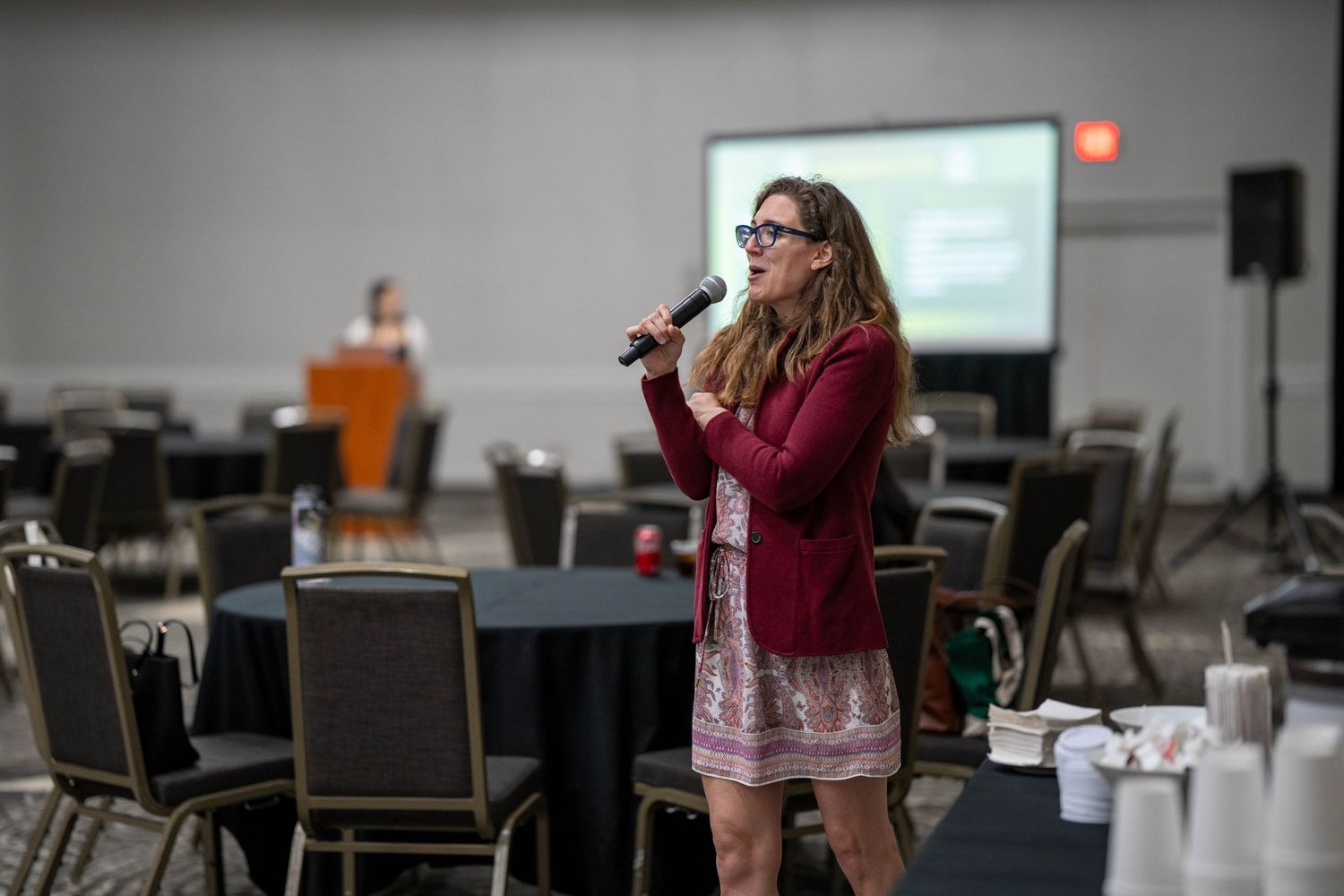 Woman in red blazer speaking into microphone at a conference event in a large banquet hall.