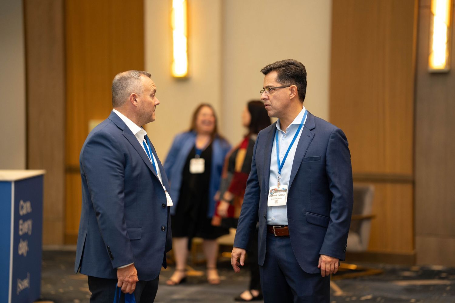 Two men in blue suits converse at a professional conference event with attendees visible in the background.