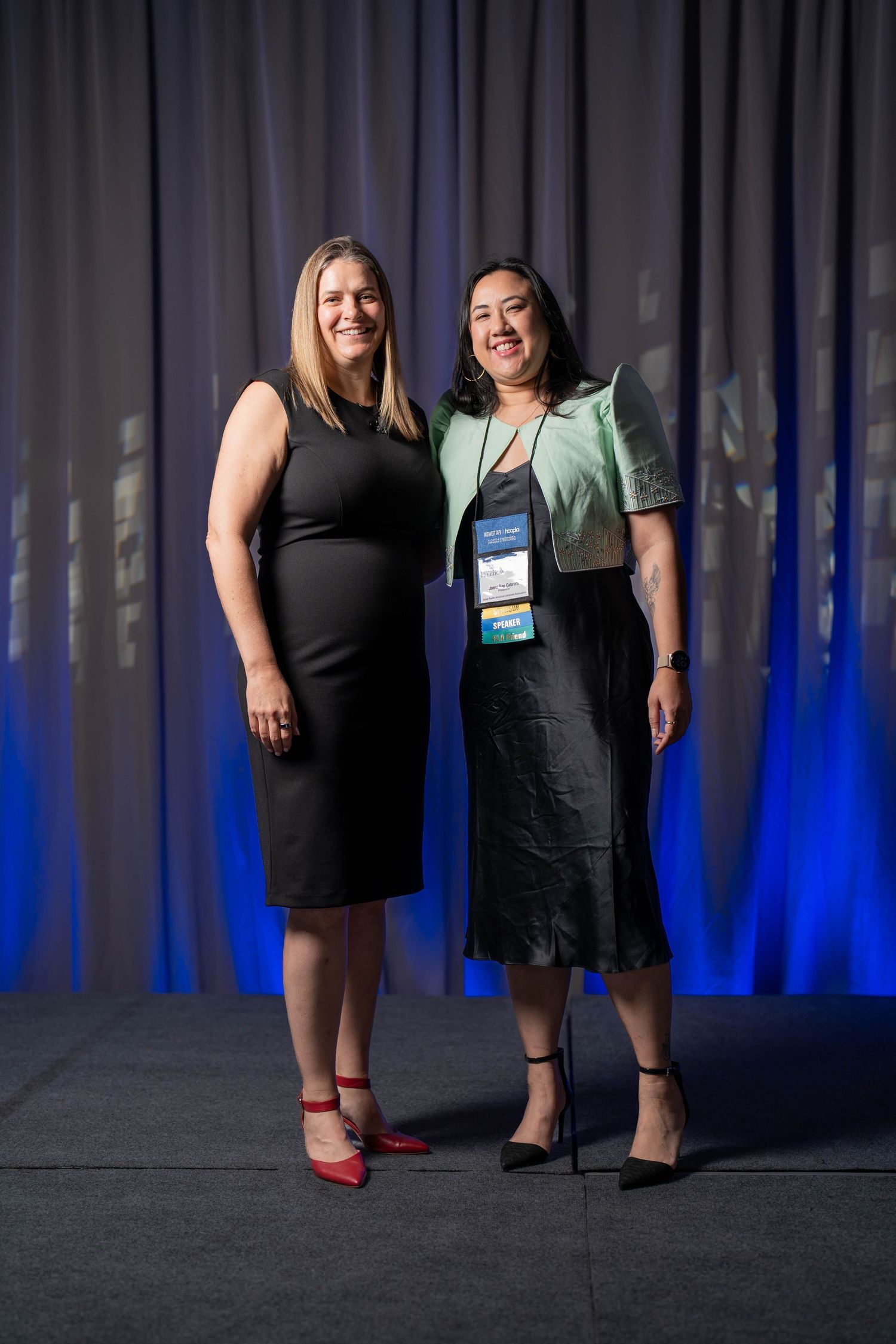 Two women in formal attire smile together on stage at a professional conference event.