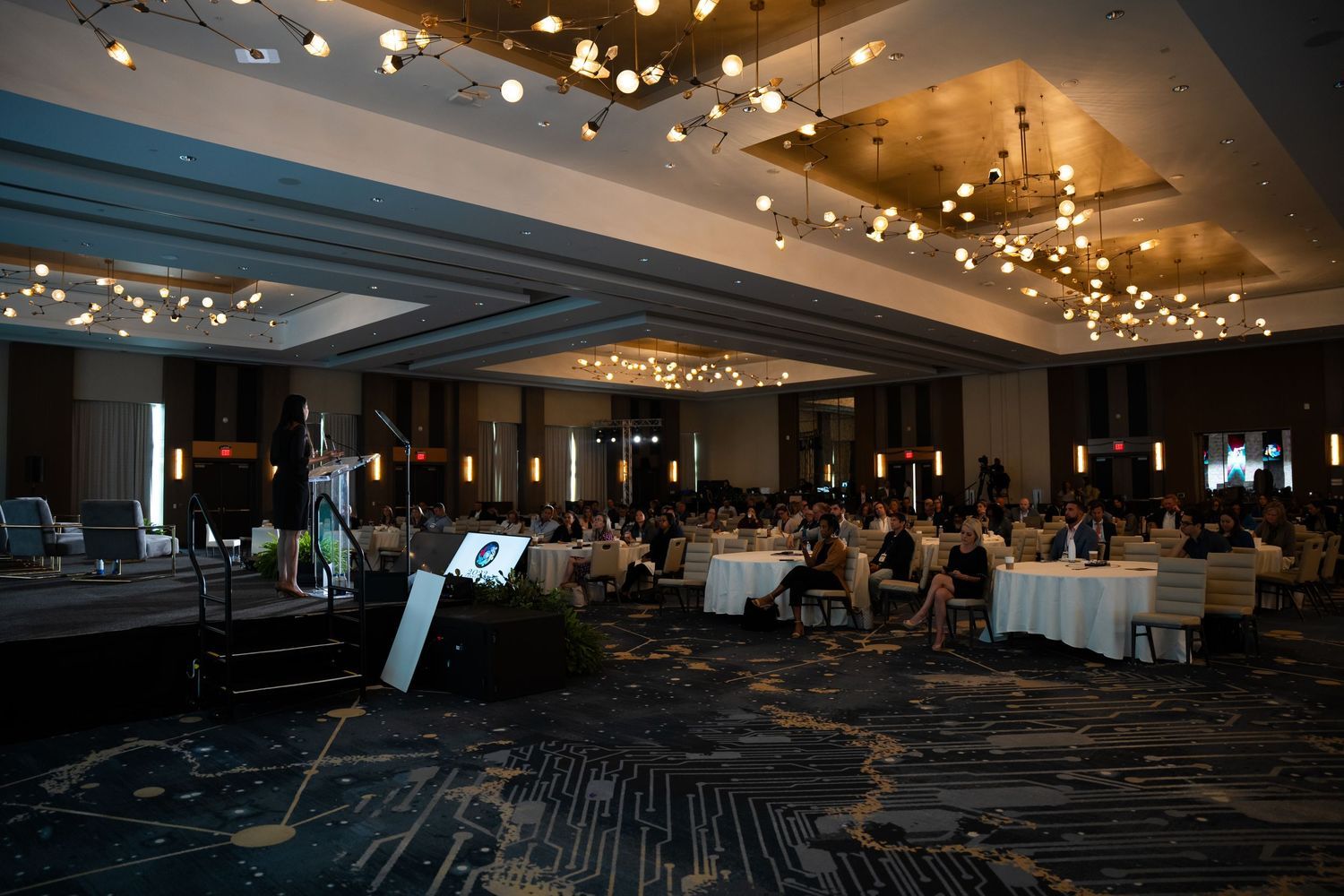 Elegant hotel ballroom with string lights, round dining tables, and guests gathered for a formal event.