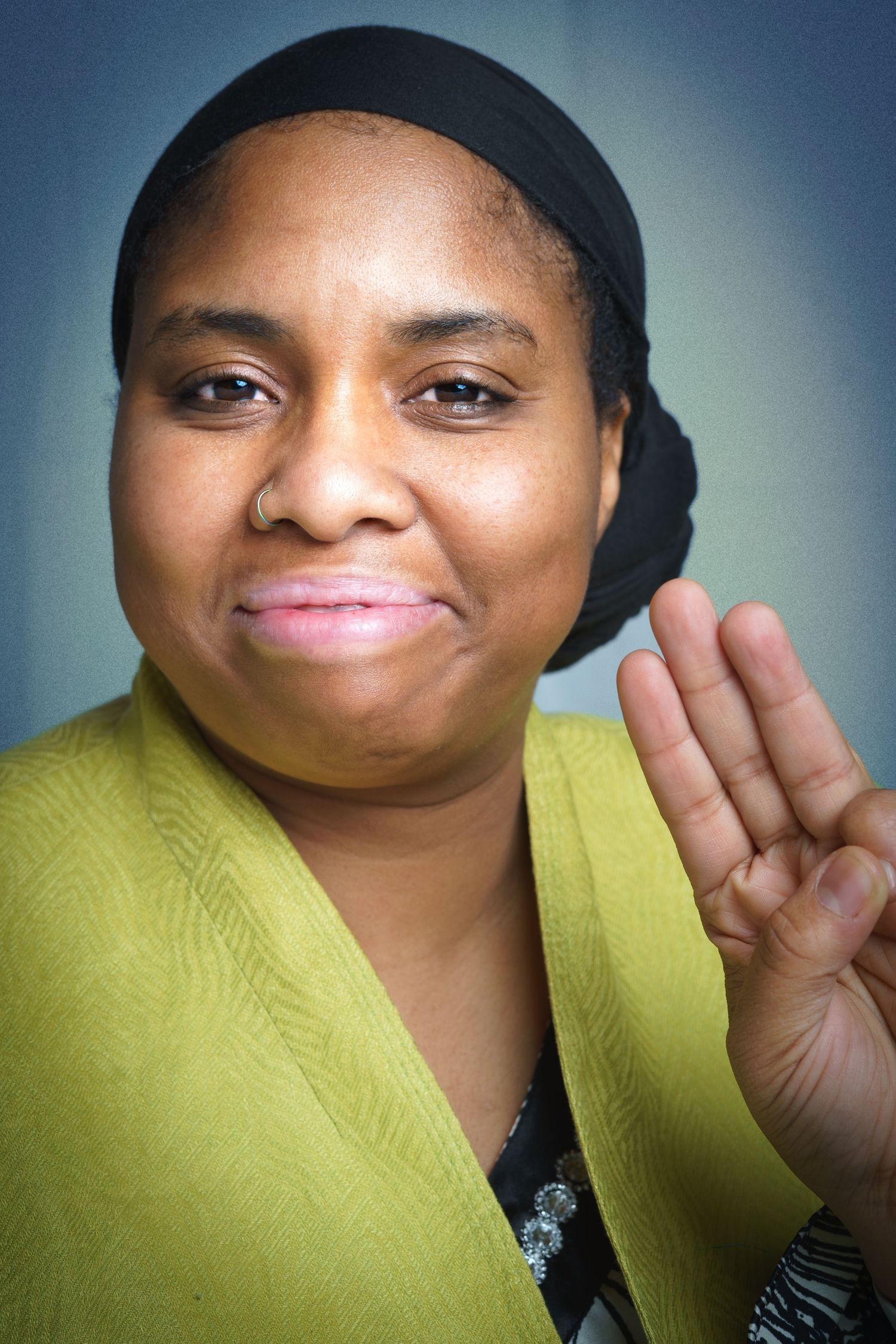 Close-up portrait photograph of a woman looking directly at the camera, holding up three fingers.