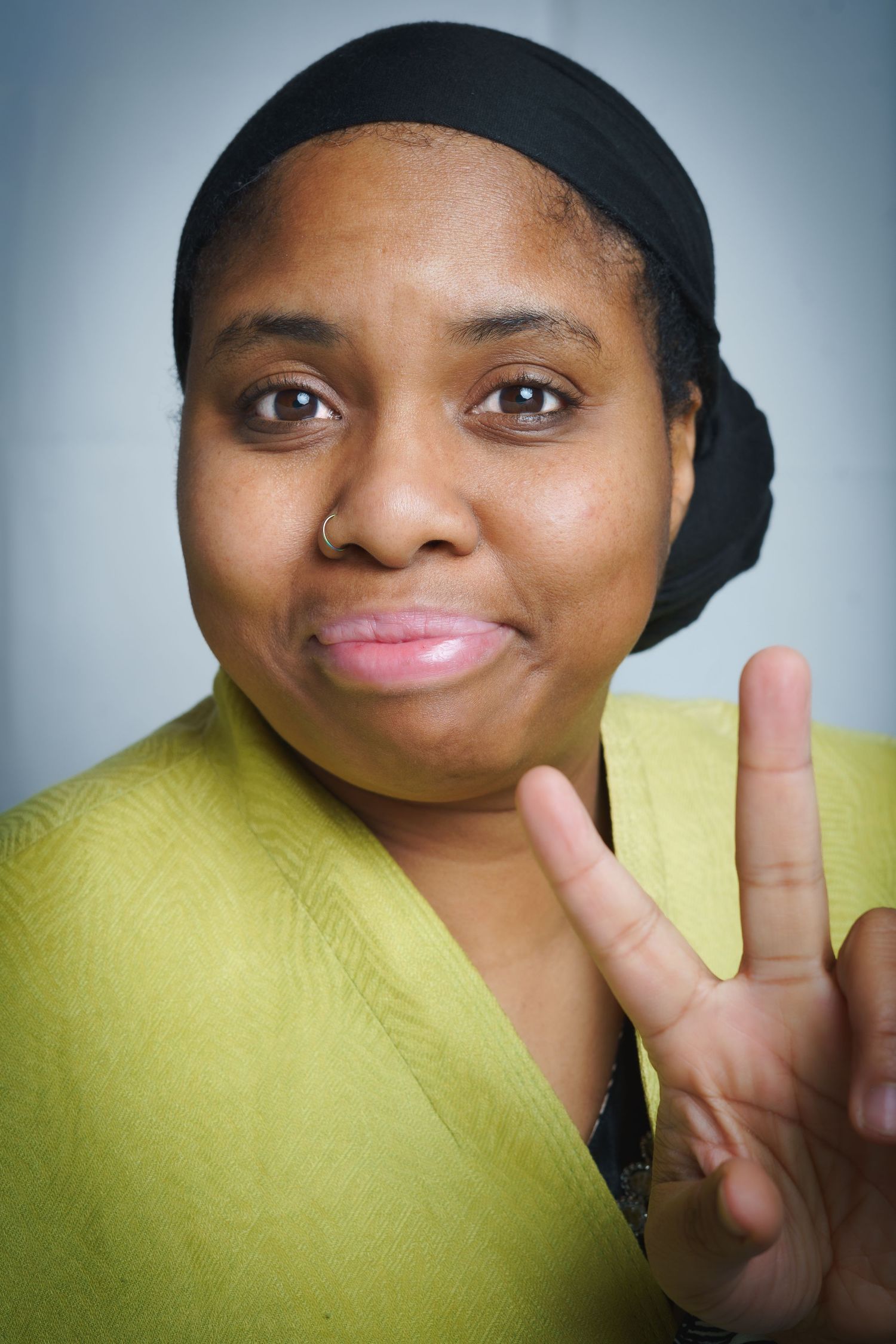 Close-up portrait photograph of a woman looking directly at the camera, holding up two fingers.