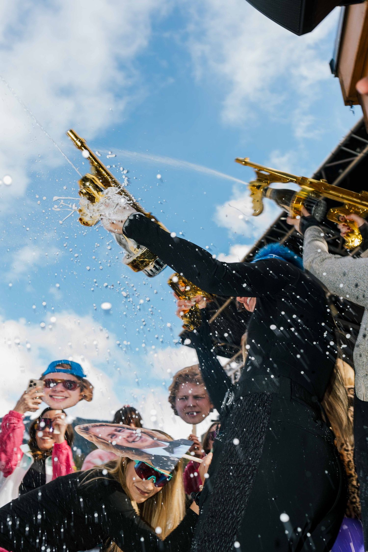 Person spraying champagne from a gold bottle at a lively outdoor ski resort festival.