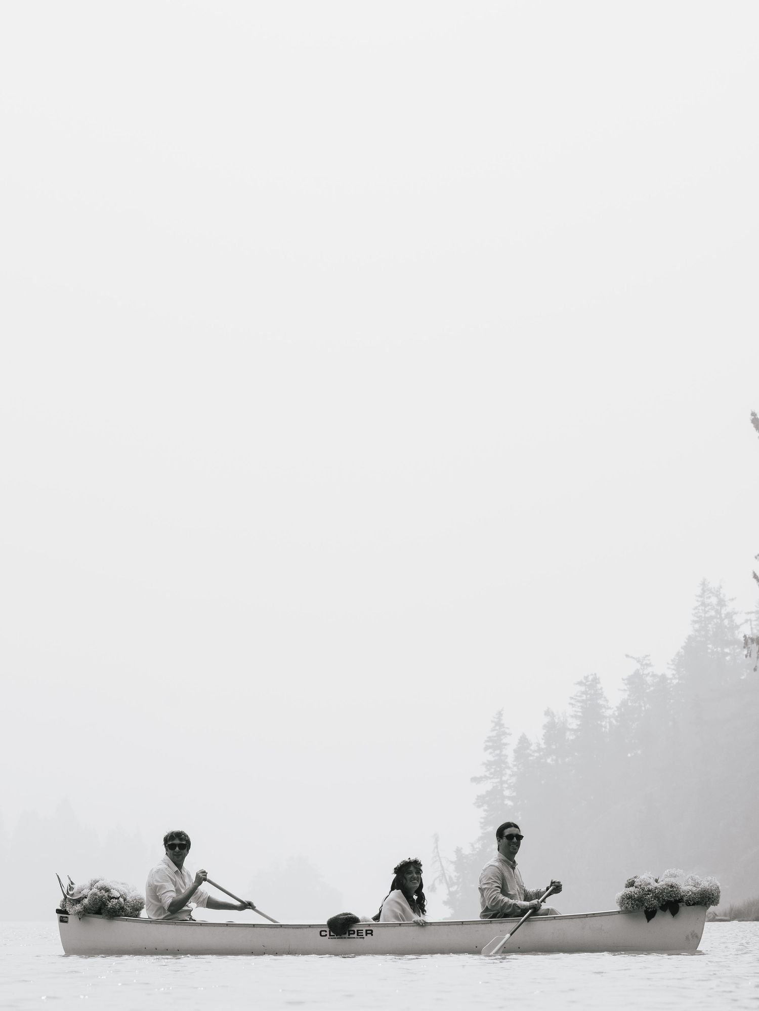 Three people paddling a canoe through misty foggy lake surrounded by pine trees in black and white.