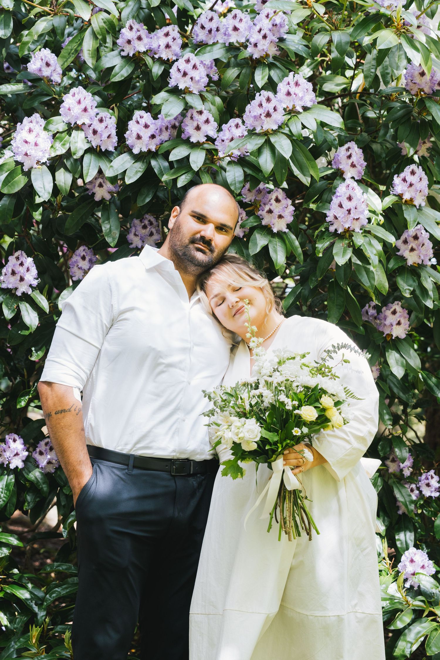 Newlywed couple in white attire posing before blooming purple rhododendron bushes with bridal bouquet.