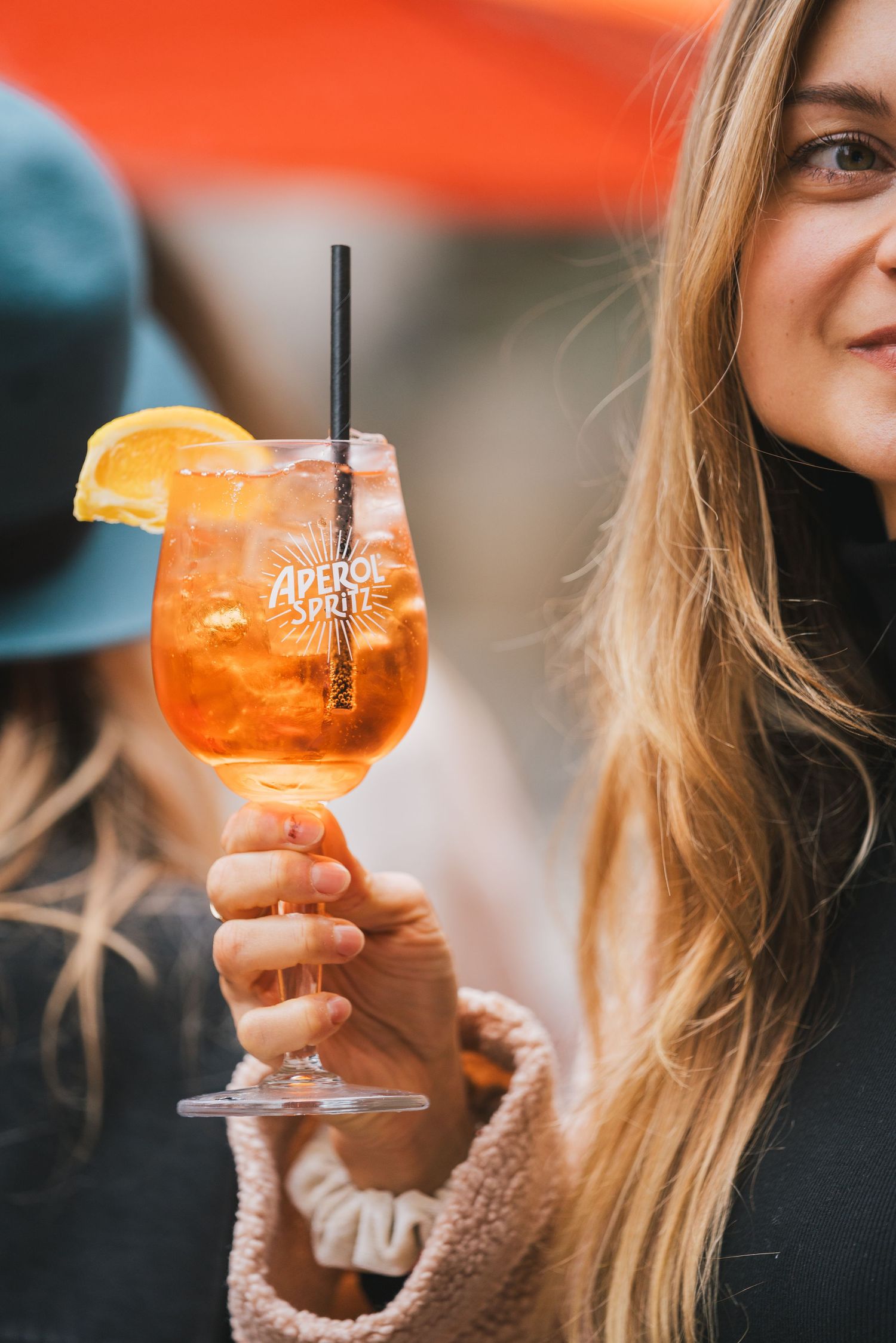 Smiling blonde woman holding an Aperol Spritz cocktail garnished with an orange slice outdoors.