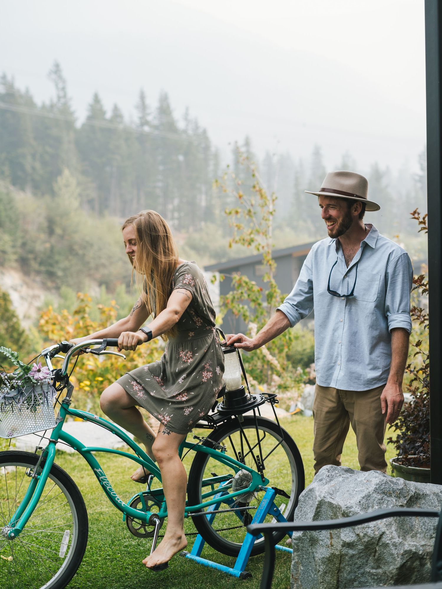 Smiling man in a hat helps a barefoot woman ride a teal cruiser bike outdoors near a misty forest.
