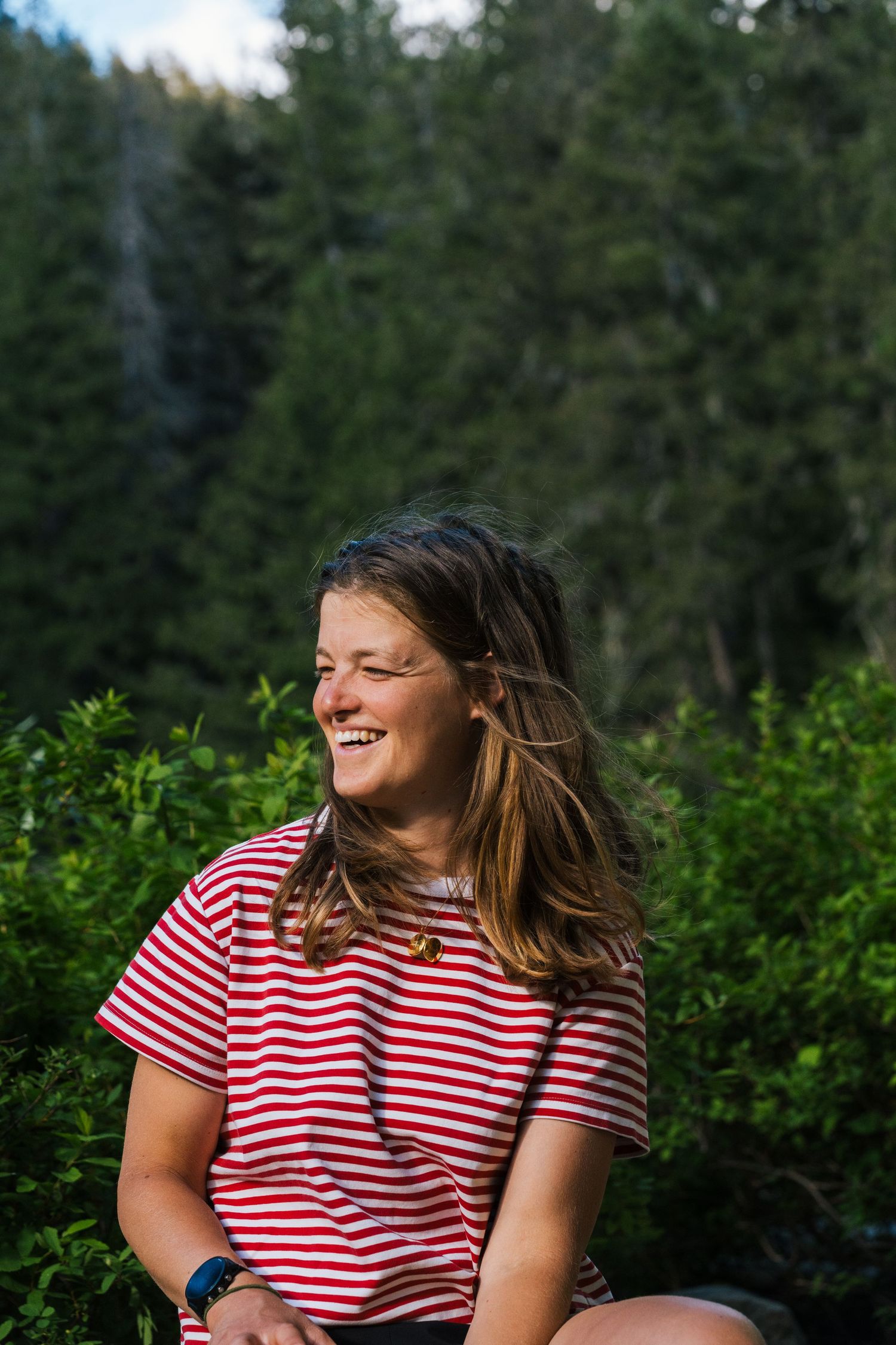 Happy young woman in red striped shirt laughing outdoors surrounded by lush green forest trees.